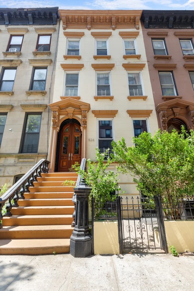 Six-story building with ornate brown and beige architectural details, front stairs leading to a wooden door, and a small gated garden area with green bushes.