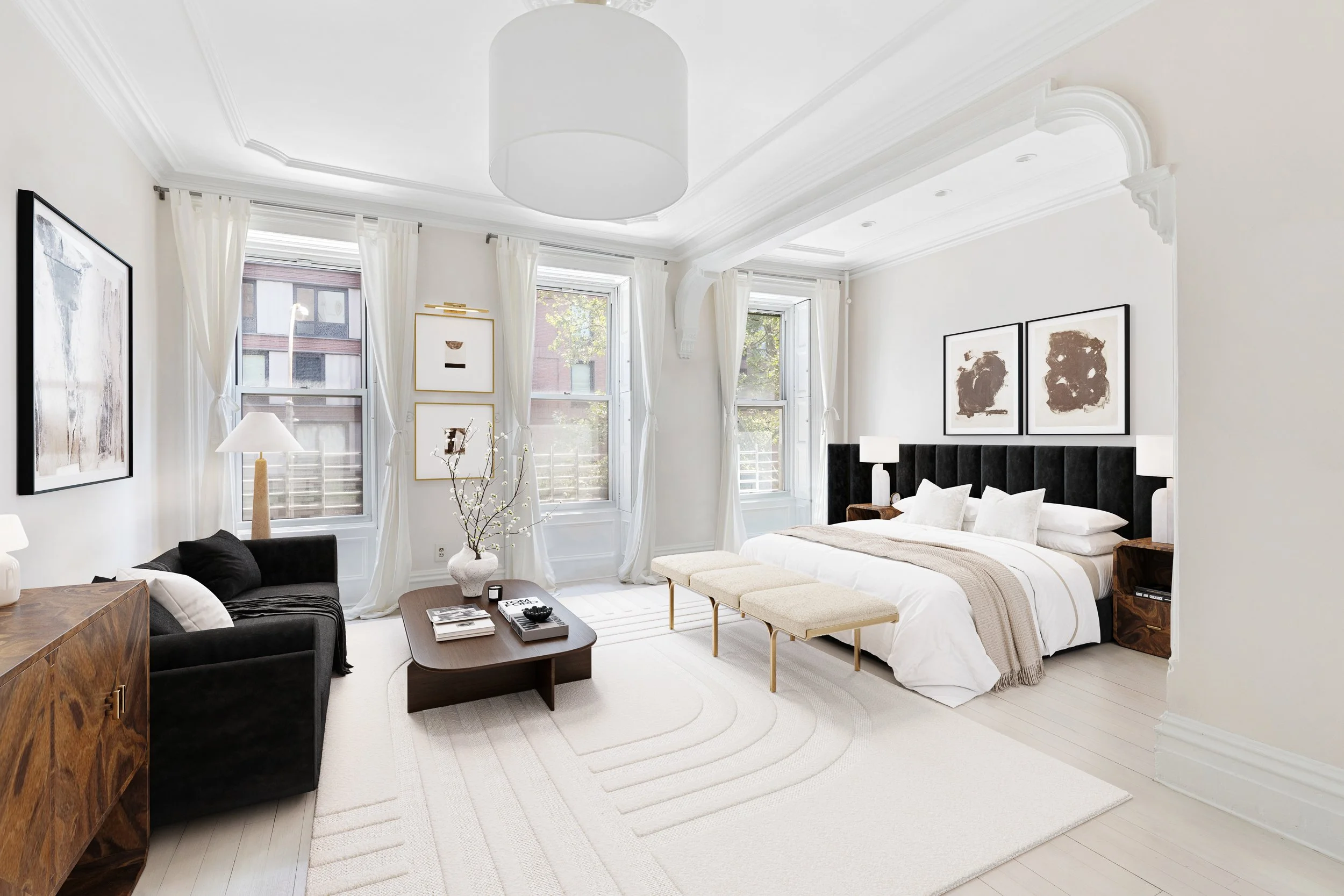 Bright and modern bedroom with large windows, white walls, and minimalist decor. Contains a bed with a black headboard, nightstands with lamps, a black sofa, a wooden coffee table, and framed artwork.