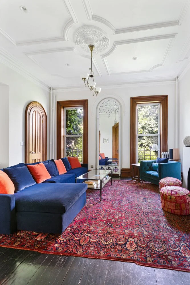 Living room with white walls, ornate ceiling, wooden trim, a dark blue sofa with orange and blue pillows, a glass coffee table, patterned area rug, two windows with wooden frames, a mirror, green armchair, and pink patterned ottomans.