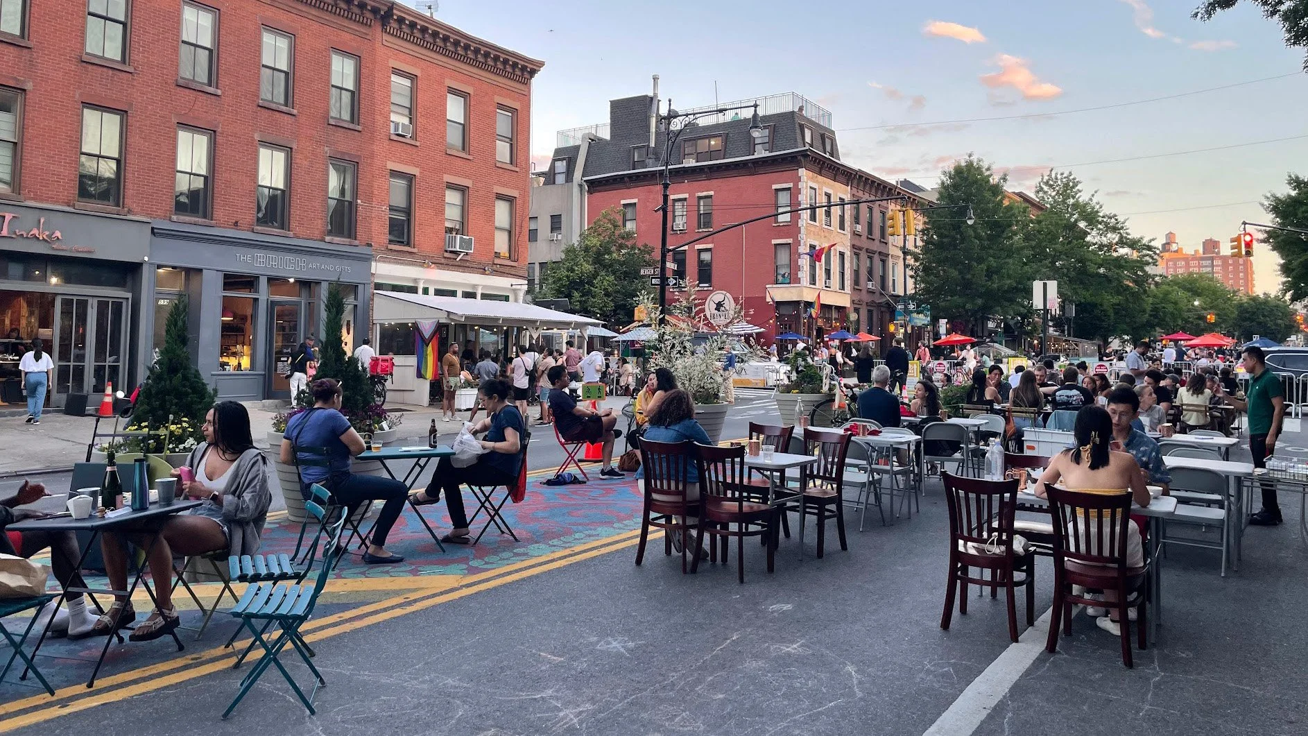 People sitting at tables and chairs on a city street during the evening, with buildings, trees, and streetlights in the background.
