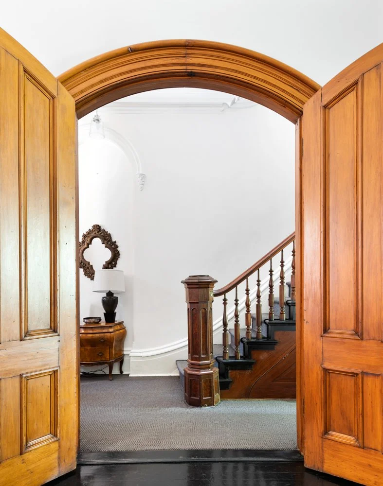 View through open wooden double doors into a hallway featuring a staircase with wooden bannister, a small wooden side table with a lamp, mirror, and decorative items, and a white wall with intricate molding.