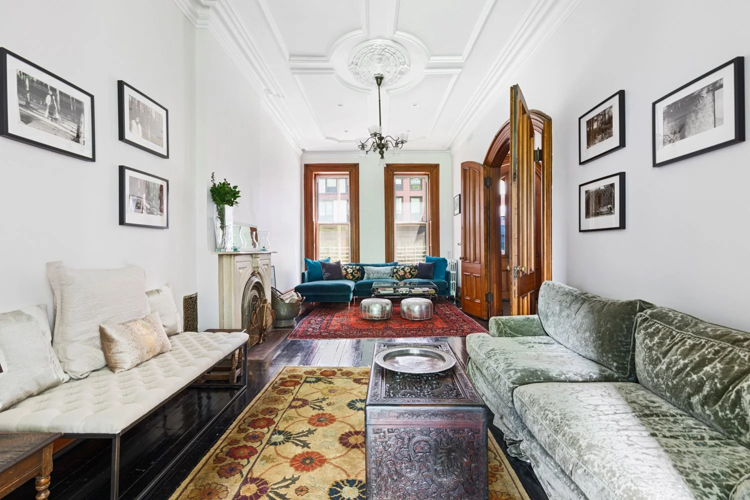 Living room with vintage and colorful furniture, including a green velvet sofa, white cushioned bench, blue velvet couch, and ornate wooden furniture, with framed black-and-white photos on the white walls, hardwood floors, and rugs, natural light from large windows, and a chandelier on the ceiling.