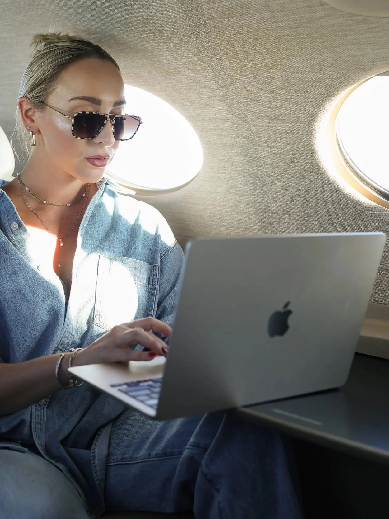 A woman wearing sunglasses and a denim shirt is sitting inside an airplane, working on a MacBook with the airplane window in the background.