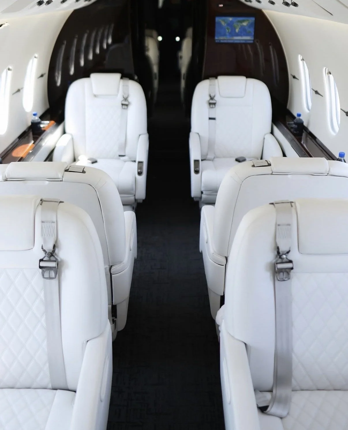 Interior of a private jet with white leather seats, some equipped with harnesses, and a small screen on the wall displaying a world map.