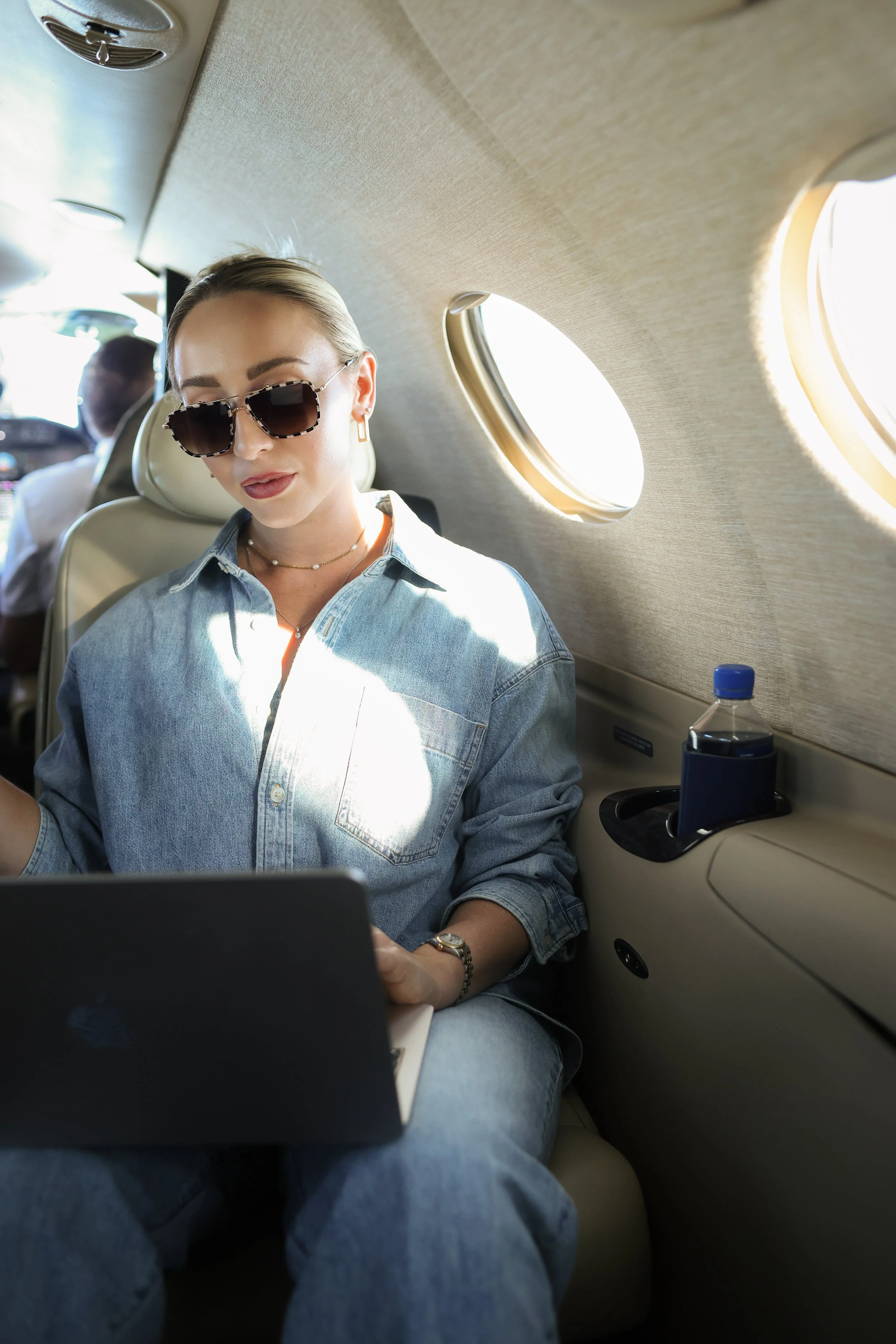 A woman with blonde hair wearing sunglasses, a denim shirt, and jewelry, sitting inside a private jet and looking at a laptop, with sunlight coming through oval windows.