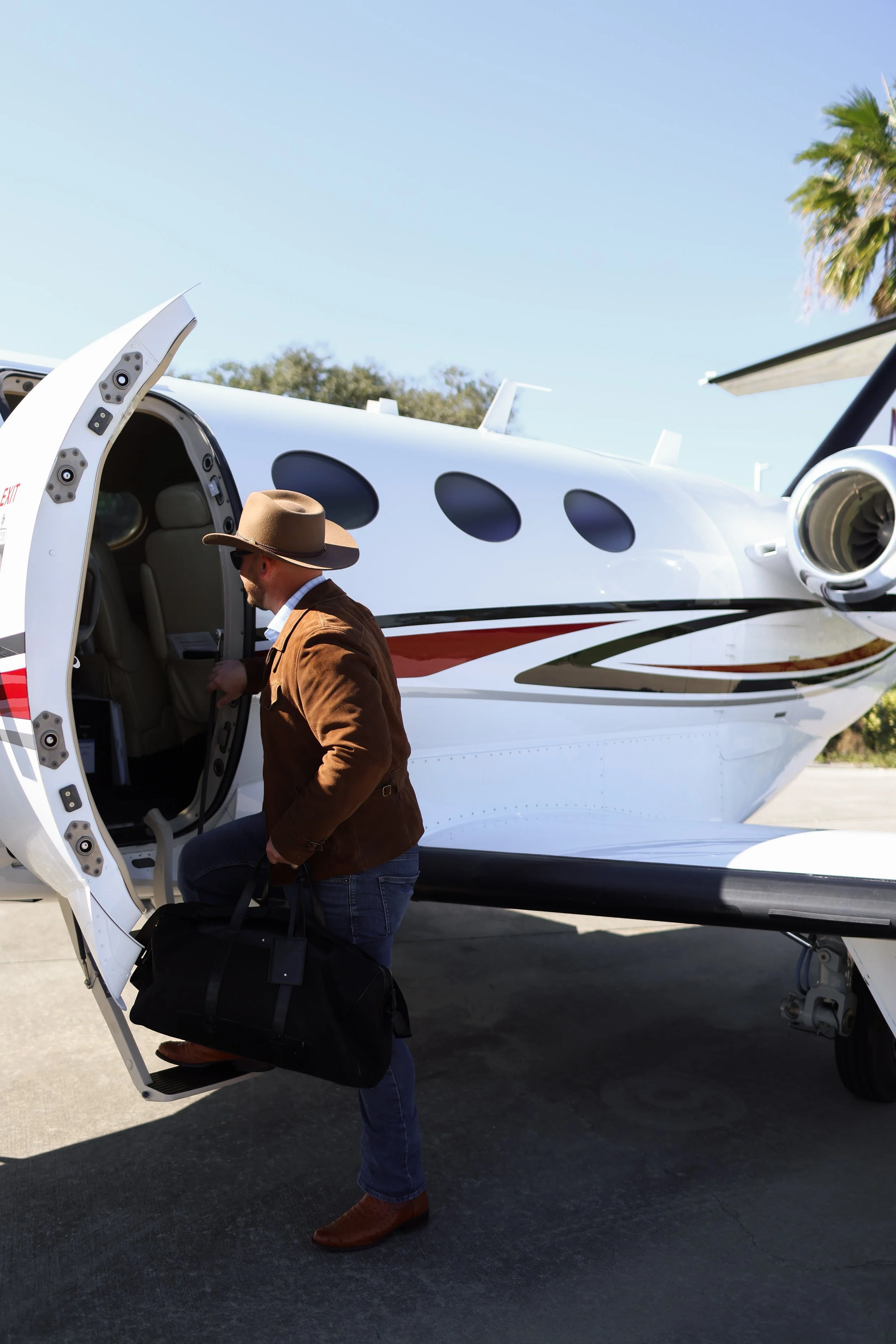 A man wearing a brown jacket, jeans, a wide-brimmed hat, and brown shoes is boarding a private jet, carrying a black duffel bag, on a clear day.