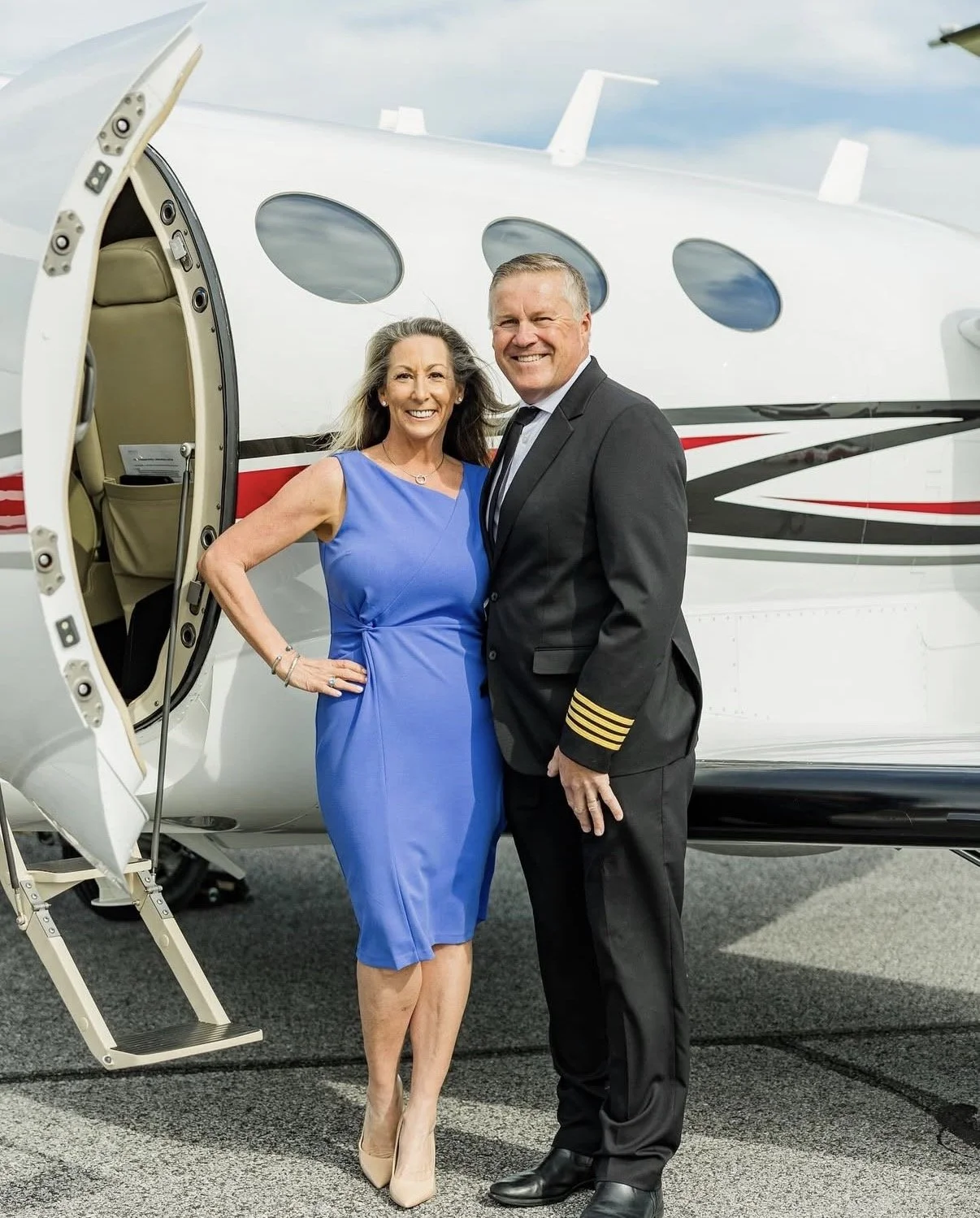 A smiling woman in a blue dress and a man in a pilot's uniform stand in front of a private jet, posing for the photo.