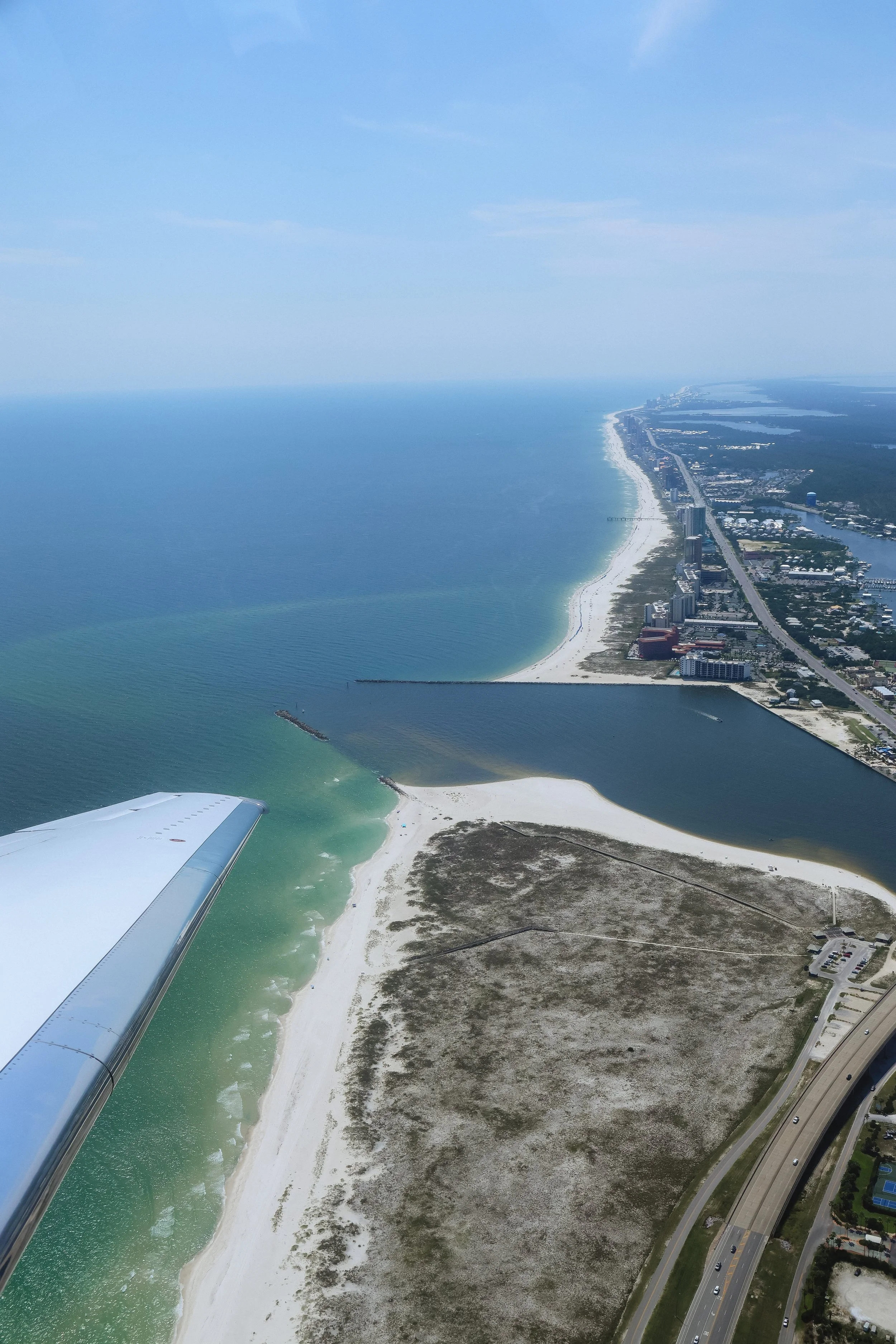 Aerial view of a beach coastline with a clear blue sky, visible ocean, sandy beach, and a coastal city with tall buildings along the shore. Part of an airplane wing is visible in the foreground.