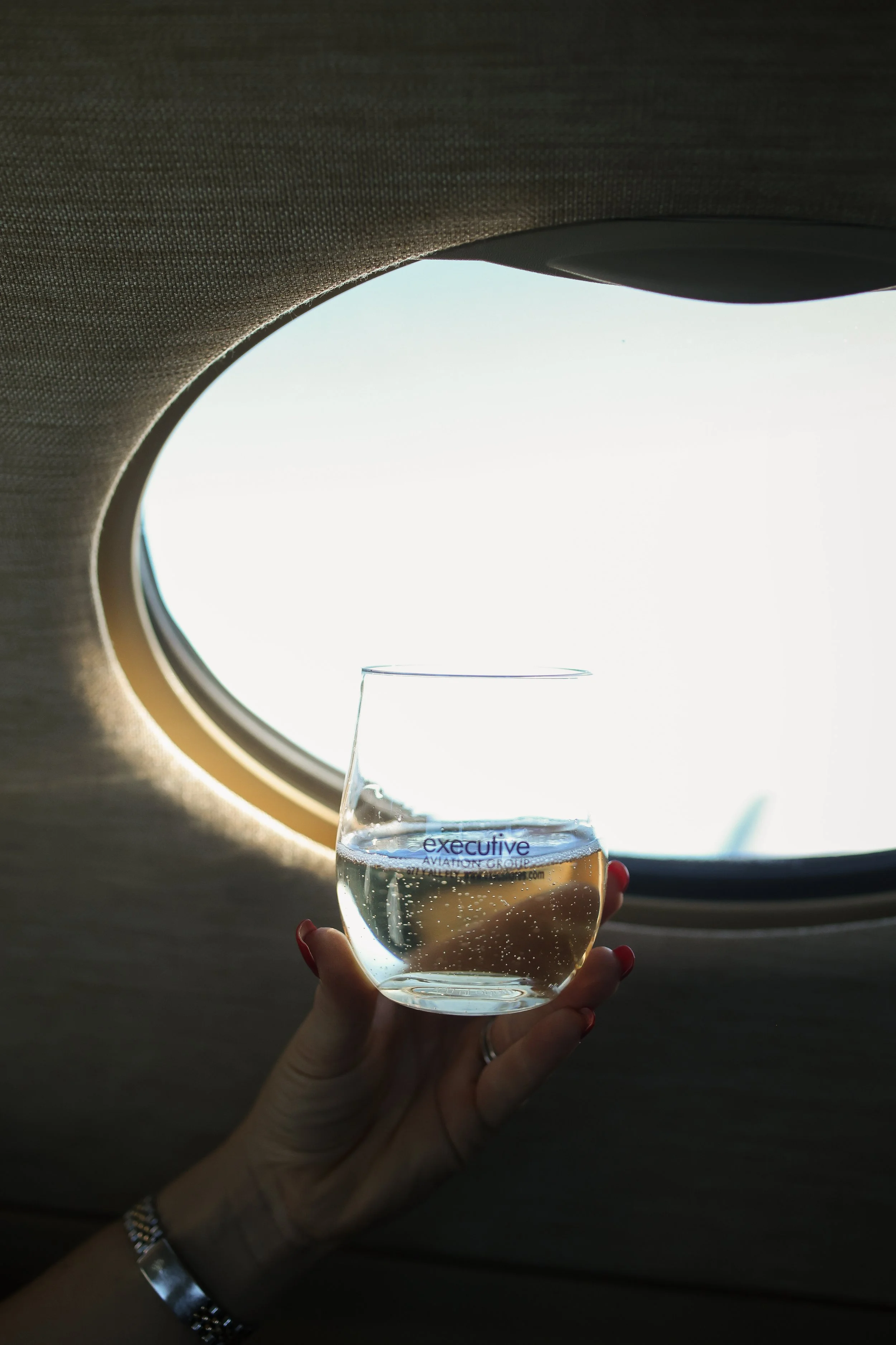 A hand holding a glass of sparkling water inside an airplane cabin near a small oval window, with a bright sky outside.