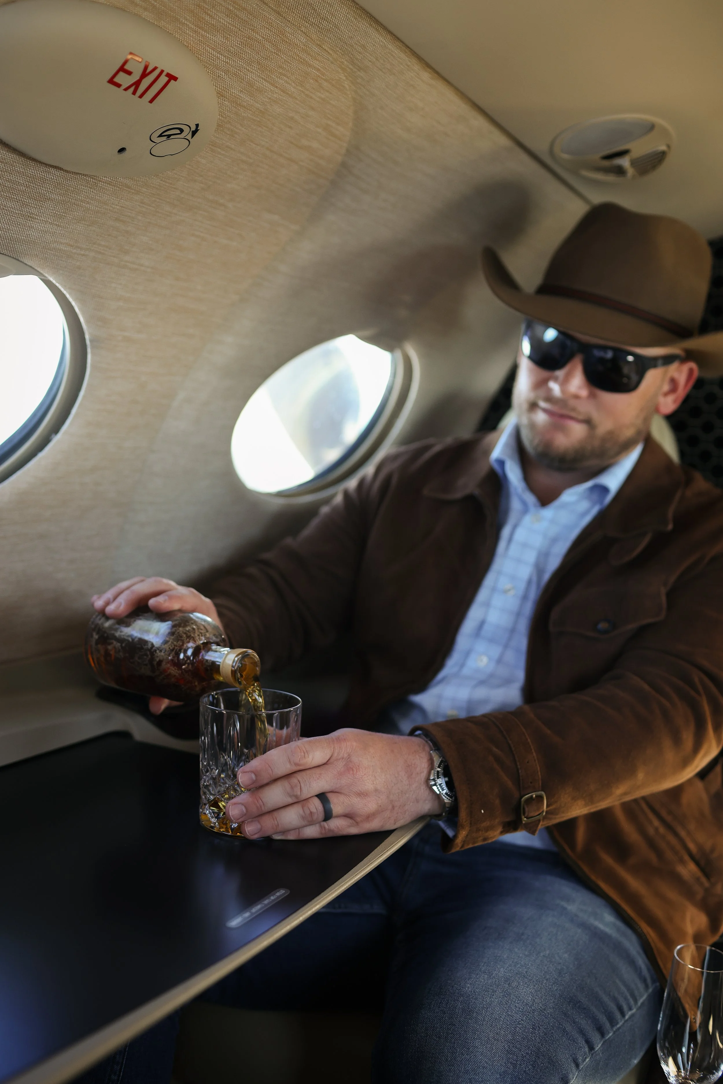 A man wearing sunglasses, a cowboy hat, a brown jacket, and a blue shirt, sitting inside an airplane, pouring a drink into a glass.