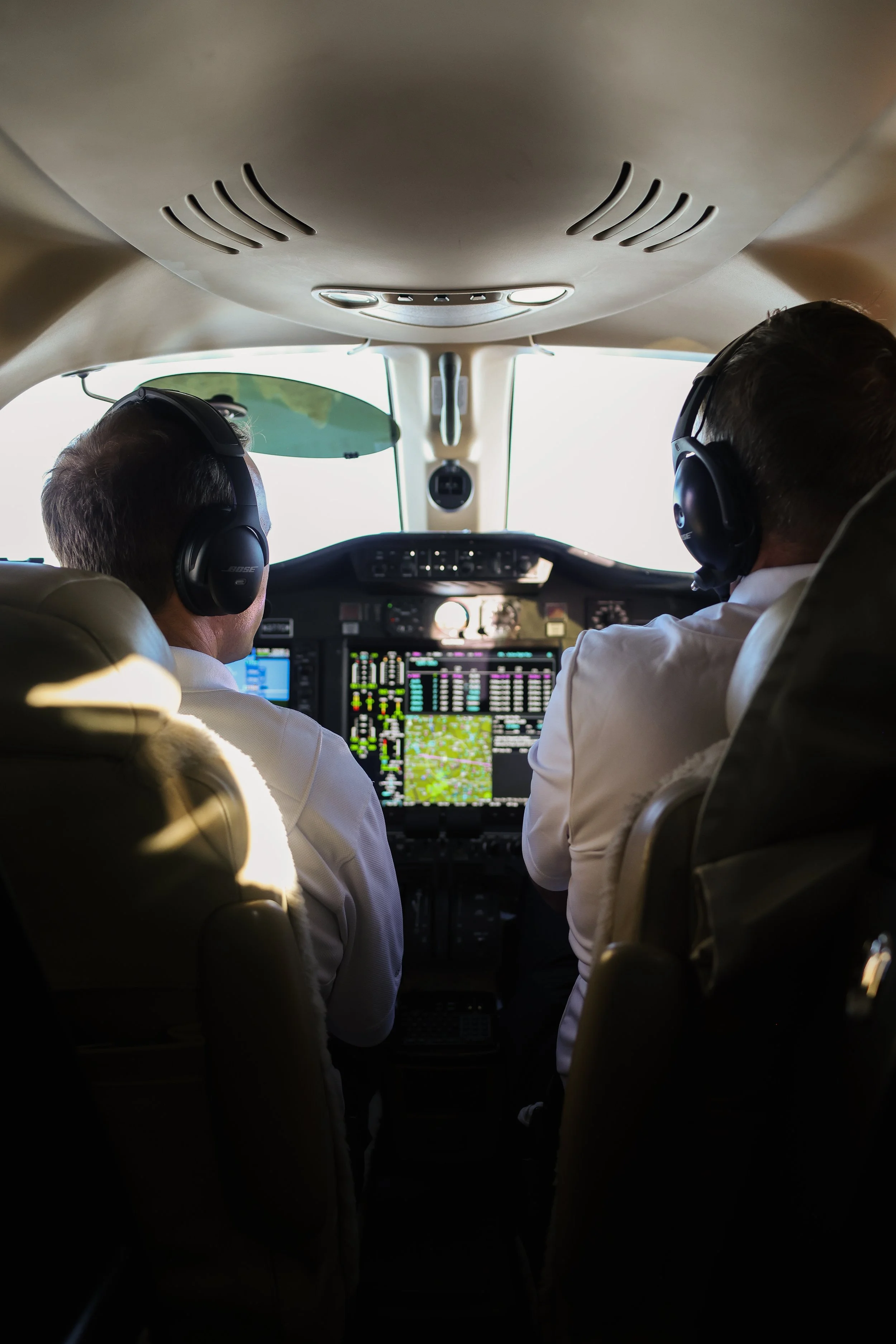 View inside the cockpit of an airplane with two pilots wearing headsets, facing the controls and screens.