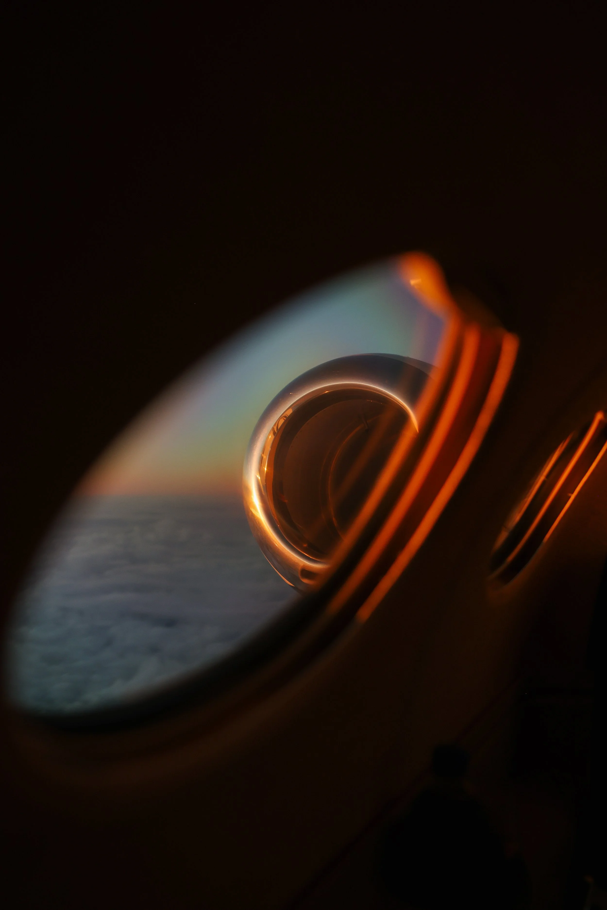 View of the airplane engine seen through the circular window at sunset with a rainbow-colored sky and clouds below.