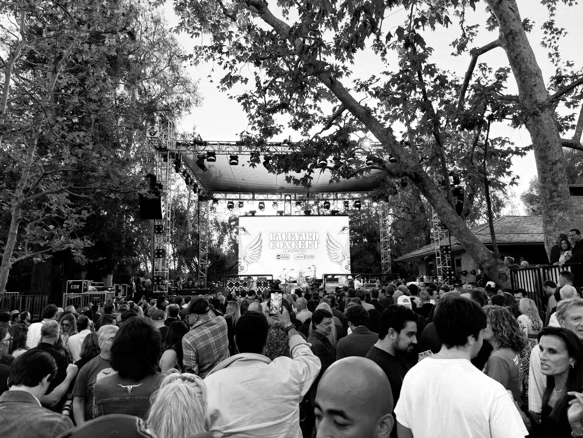 Crowd attending an outdoor concert with a large stage, trees around, and a digital screen showing 'Backyard Concert'.
