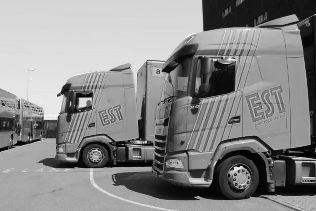 Two semi-trailer trucks parked on a road, with the letters 'EST' on the side of each truck's cabin.