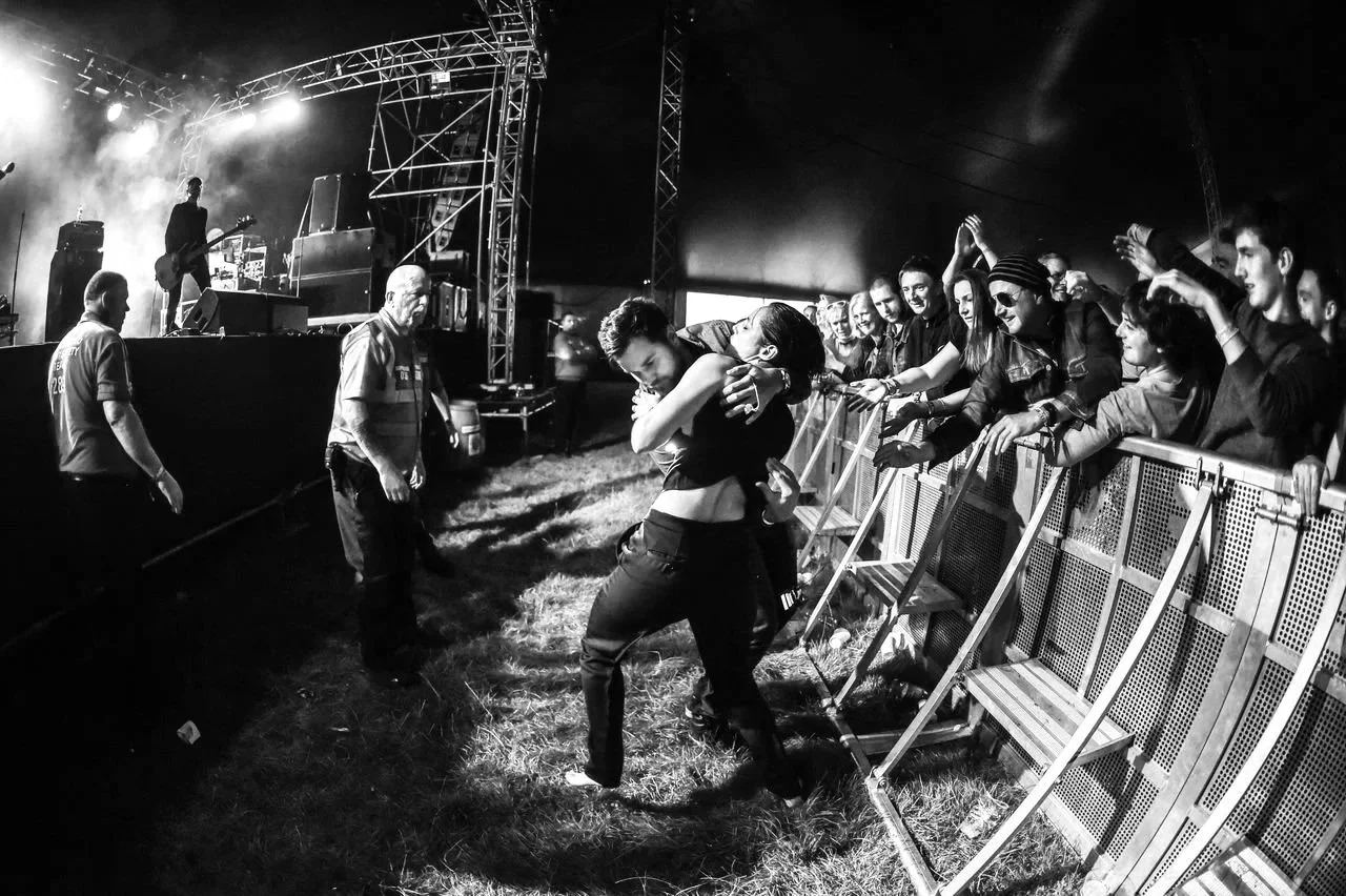 A black and white photo of a concert scene with a large audience reaching towards the stage, where a couple is hugging in front of the barrier; security guards are present by the stage.
