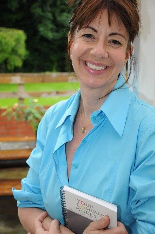 A smiling woman with short brown hair wearing a light blue button-up shirt, holding a notebook outside on a porch with a green garden background.