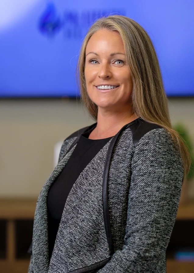 A professional woman with long blonde hair, smiling, wearing a black top and a gray blazer, standing in an office environment with a blurred screen in the background.