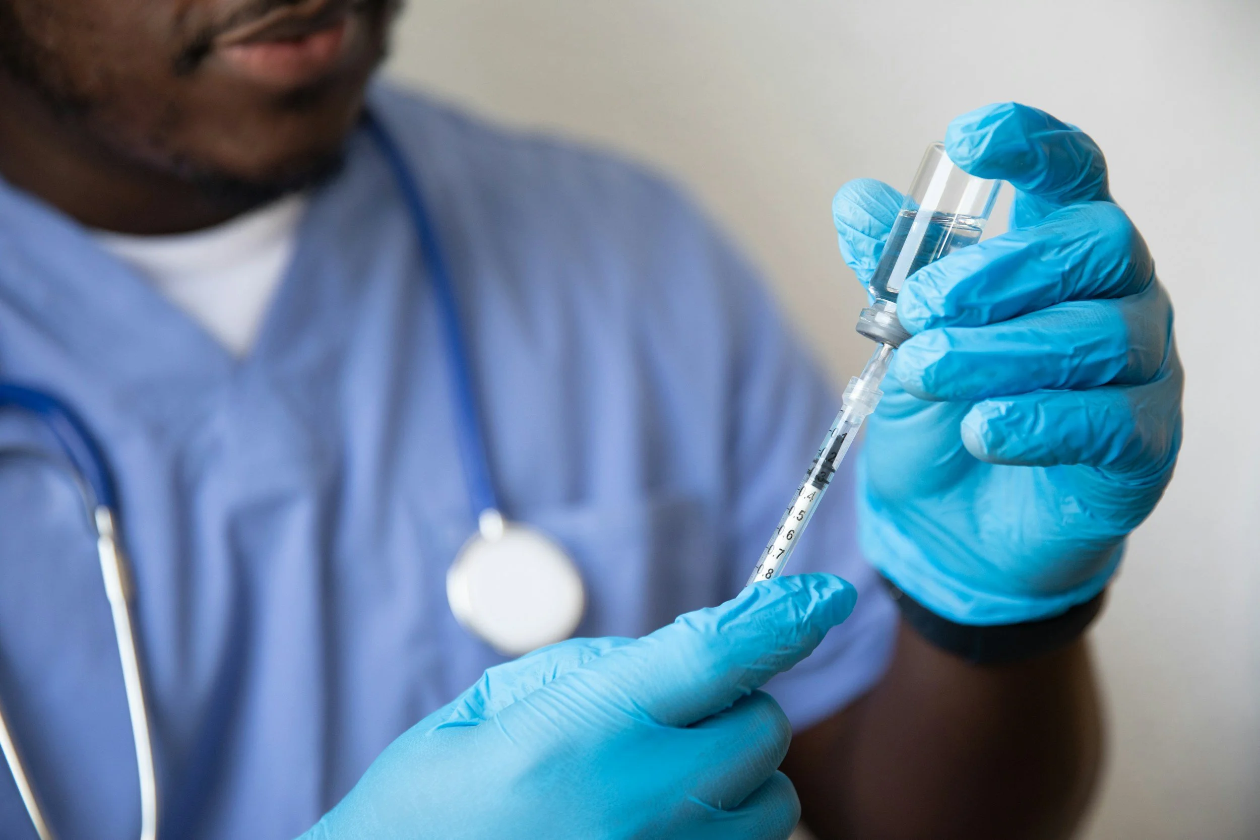 A healthcare professional in blue gloves and scrubs preparing a syringe with a vaccine or medication.