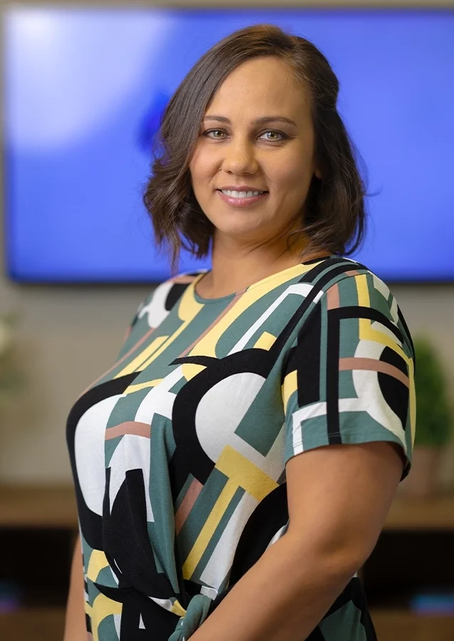 A woman with shoulder-length brown hair smiling, wearing a colorful geometric patterned dress, standing in front of a blue screen and a blurred background with plants.