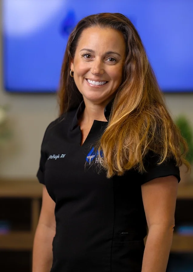 Smiling woman with long wavy brown hair wearing a black polo shirt with a logo and text, standing indoors with a blurred blue screen and plants in the background.