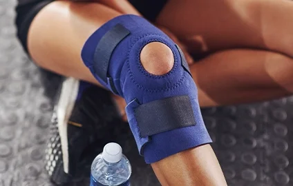 Person wearing a blue knee brace with a hole over the kneecap, sitting on the floor with a water bottle nearby.
