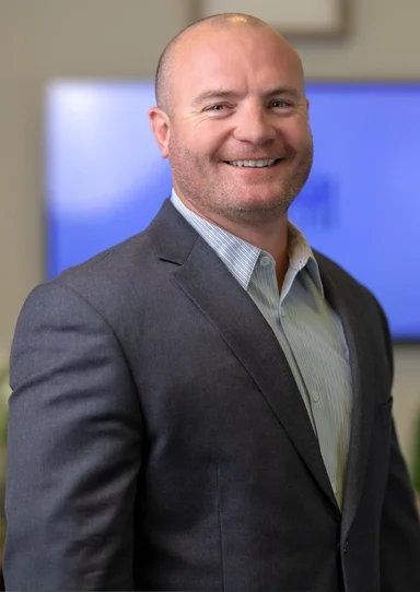A smiling man in a business suit standing indoors with a blurred television screen in the background.