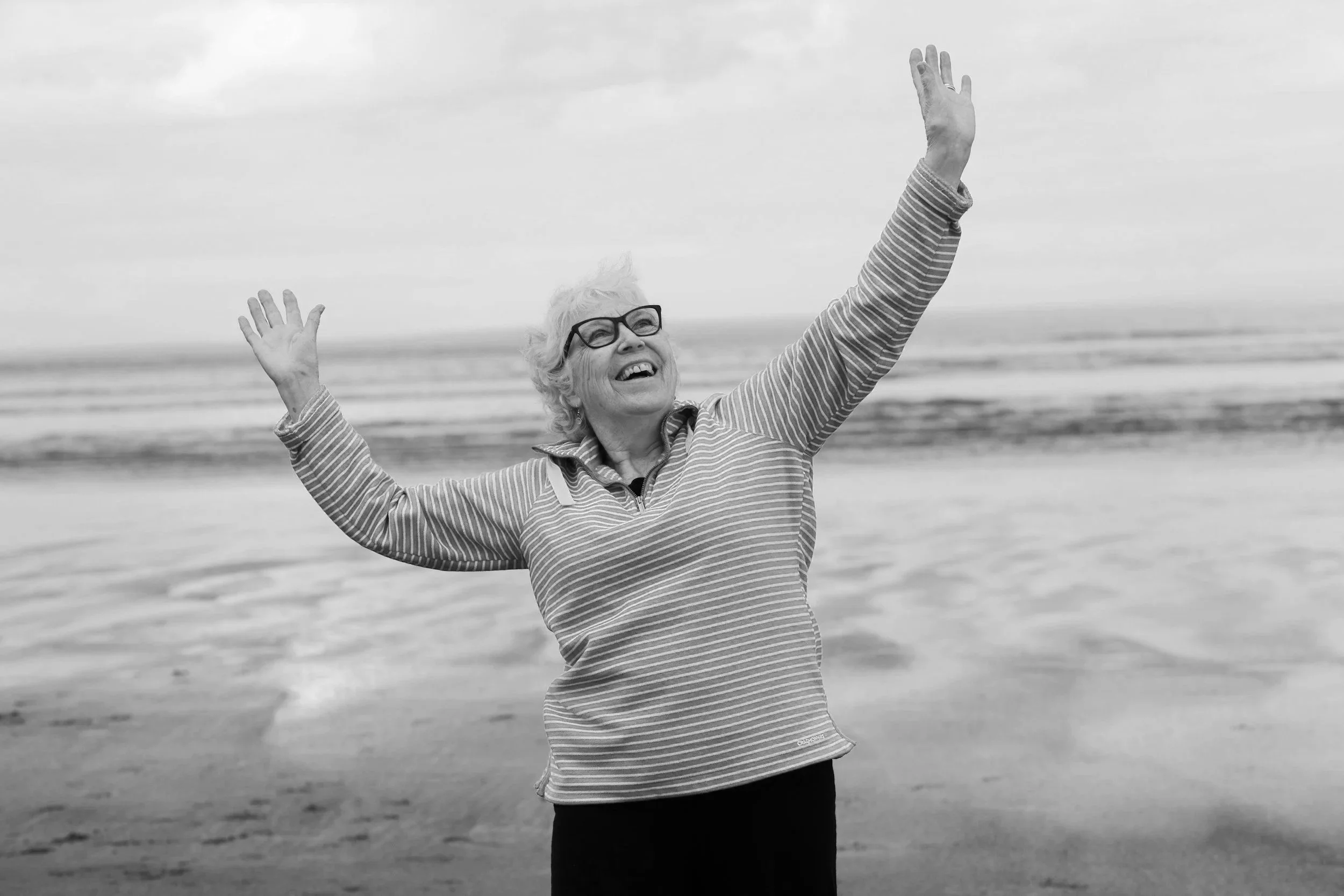 An elderly woman with glasses smiling and raising her arms at the beach.