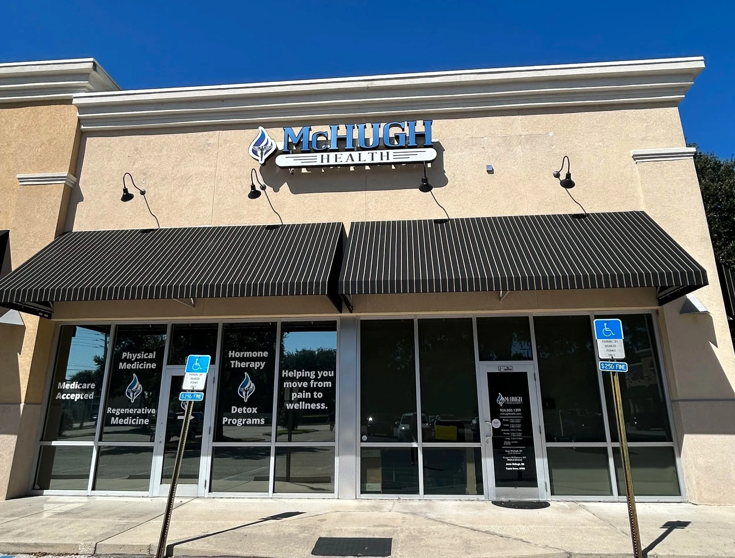 Front view of McHugh Health clinic with glass entrance doors, black-striped awnings, and a sign reading 'McHugh Health'. Two handicapped parking signs are in front of the building, and the windows display services like medical and hormone therapy.