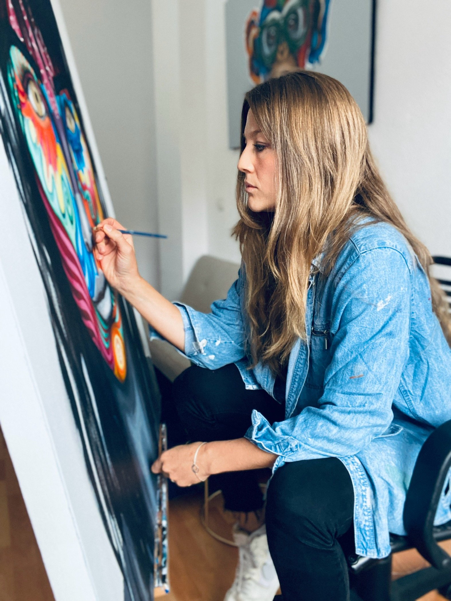 A woman with long light brown hair painting on a vertical canvas, wearing a blue denim jacket and black pants, in an indoor art studio with artwork on the wall.