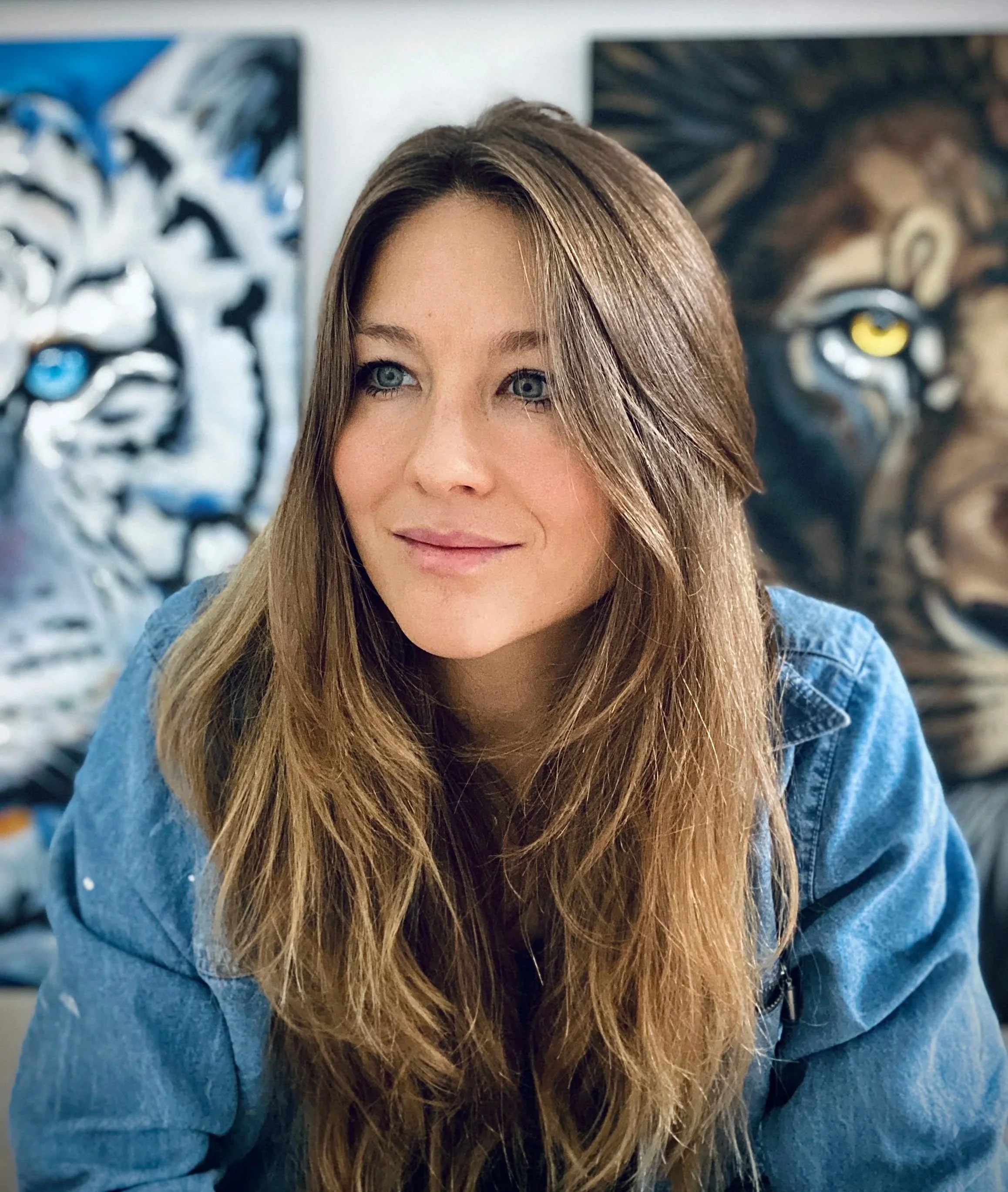 Close-up of a young woman with long brown hair, blue eyes, and fair skin, smiling slightly, wearing a blue denim jacket. In the background, there are two paintings, one abstract with blue accents and the other a realistic lion's face with a yellow eye.