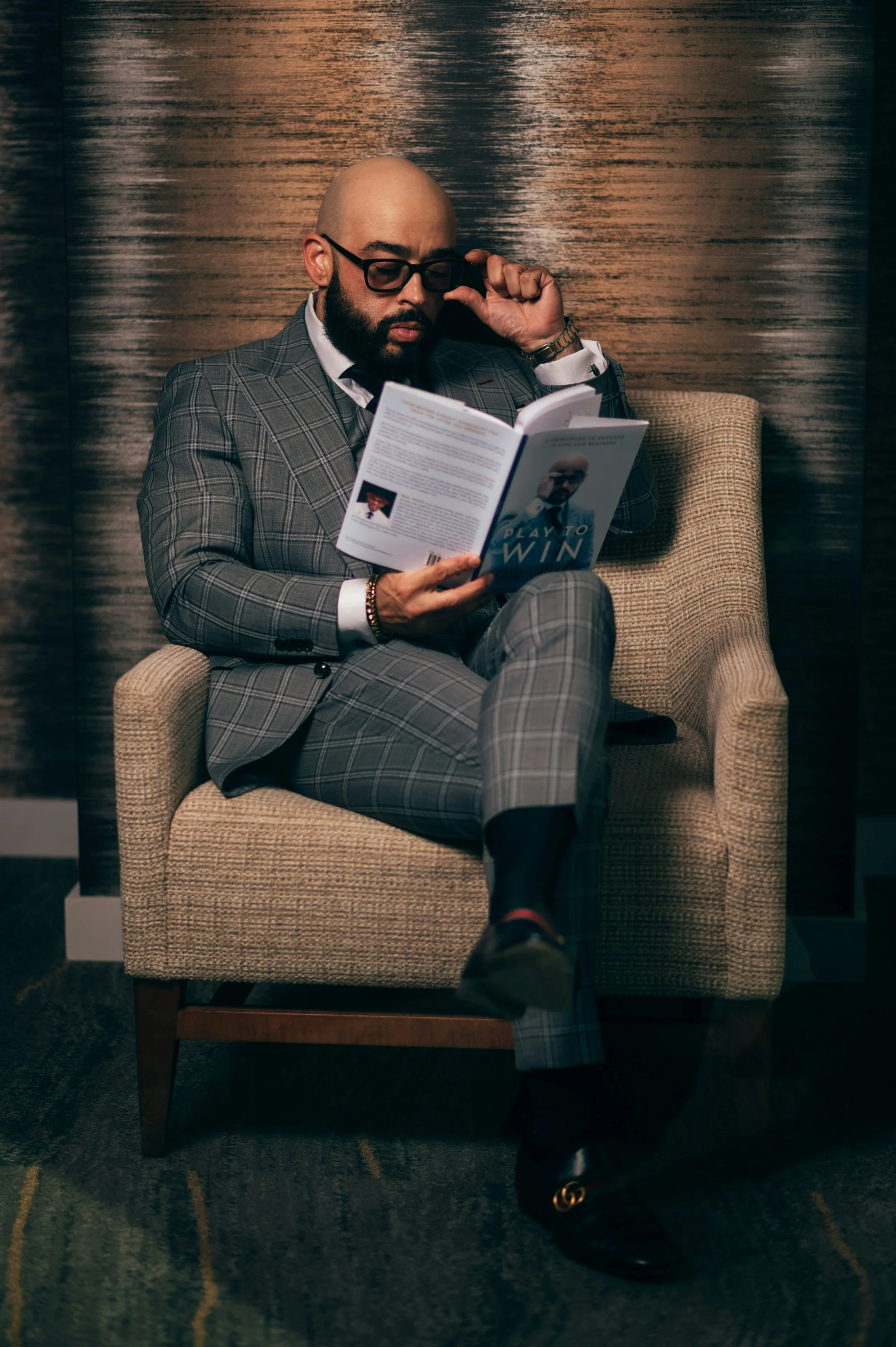 A man in a gray checkered suit with glasses reading a book while sitting in a beige armchair.