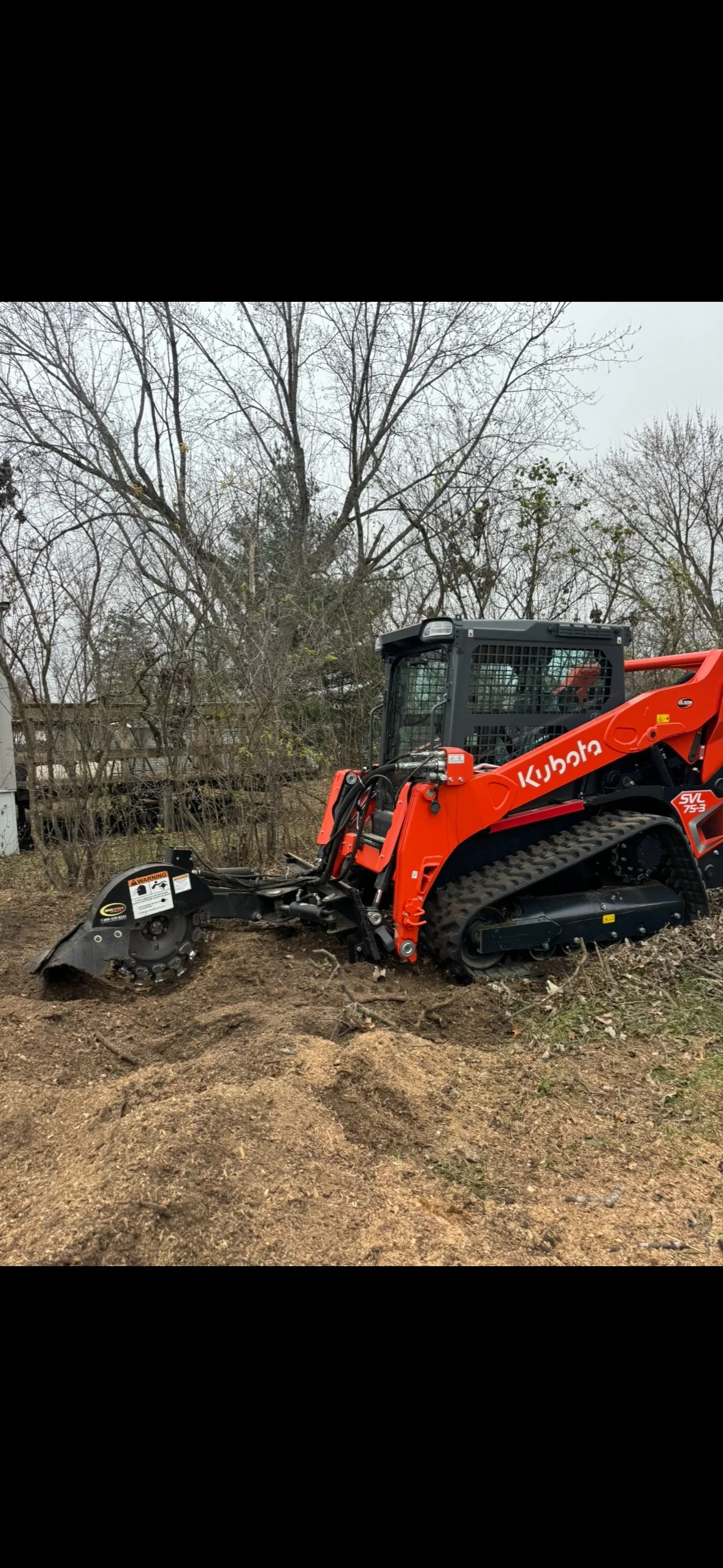 A small orange Kubota track loader with a black wheel attachment working on soil outside. Bare trees and a fence are in the background.