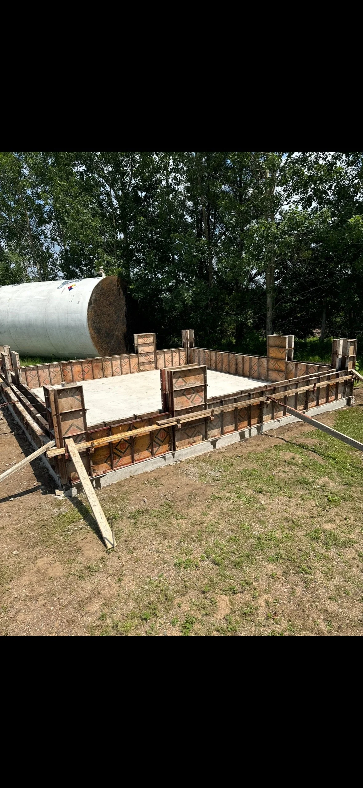 Construction site with a concrete foundation, wooden formwork for pouring walls, and a large white tank in the background, surrounded by trees.