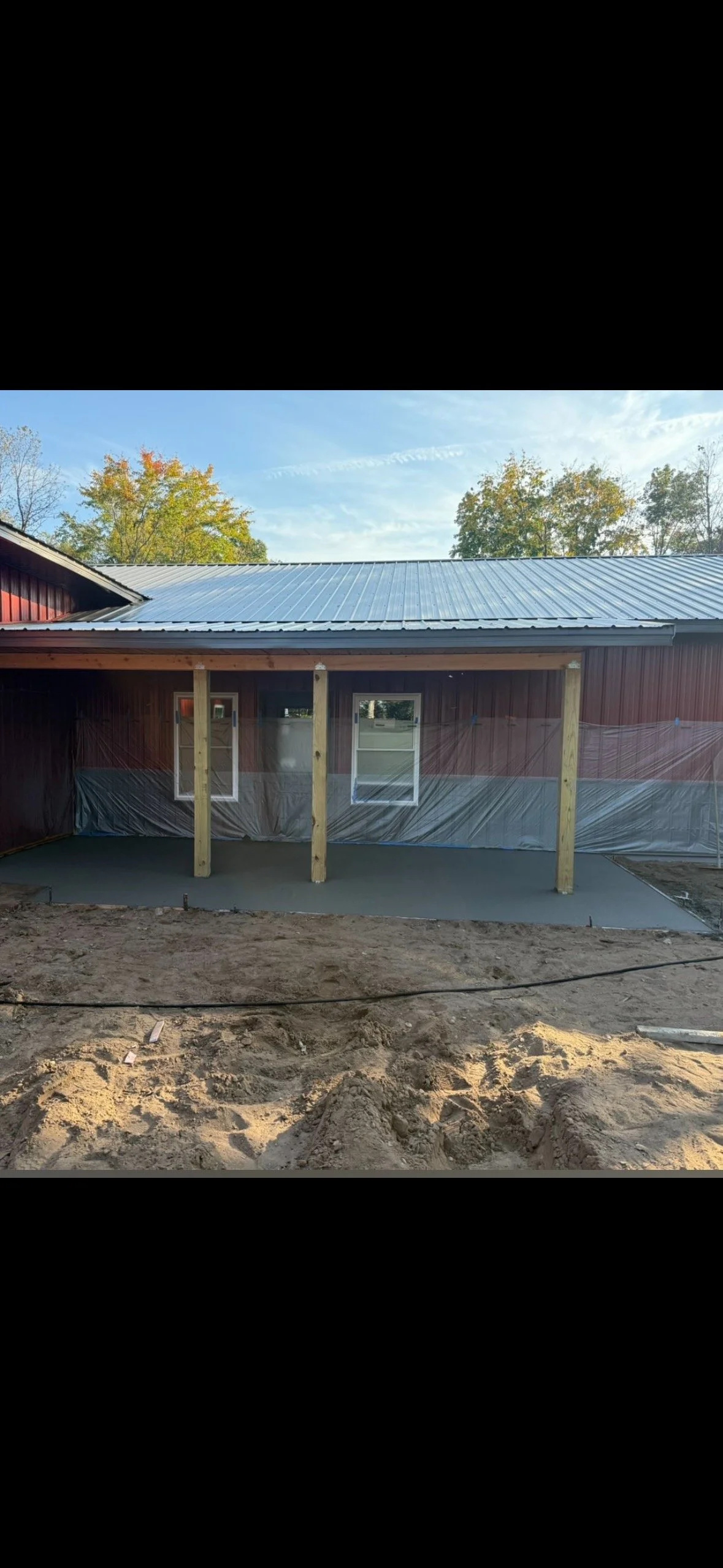 Construction site with a partially built porch, wooden support beams, and a concrete slab, attached to a red barn-style building with a metal roof, trees and blue sky in the background.