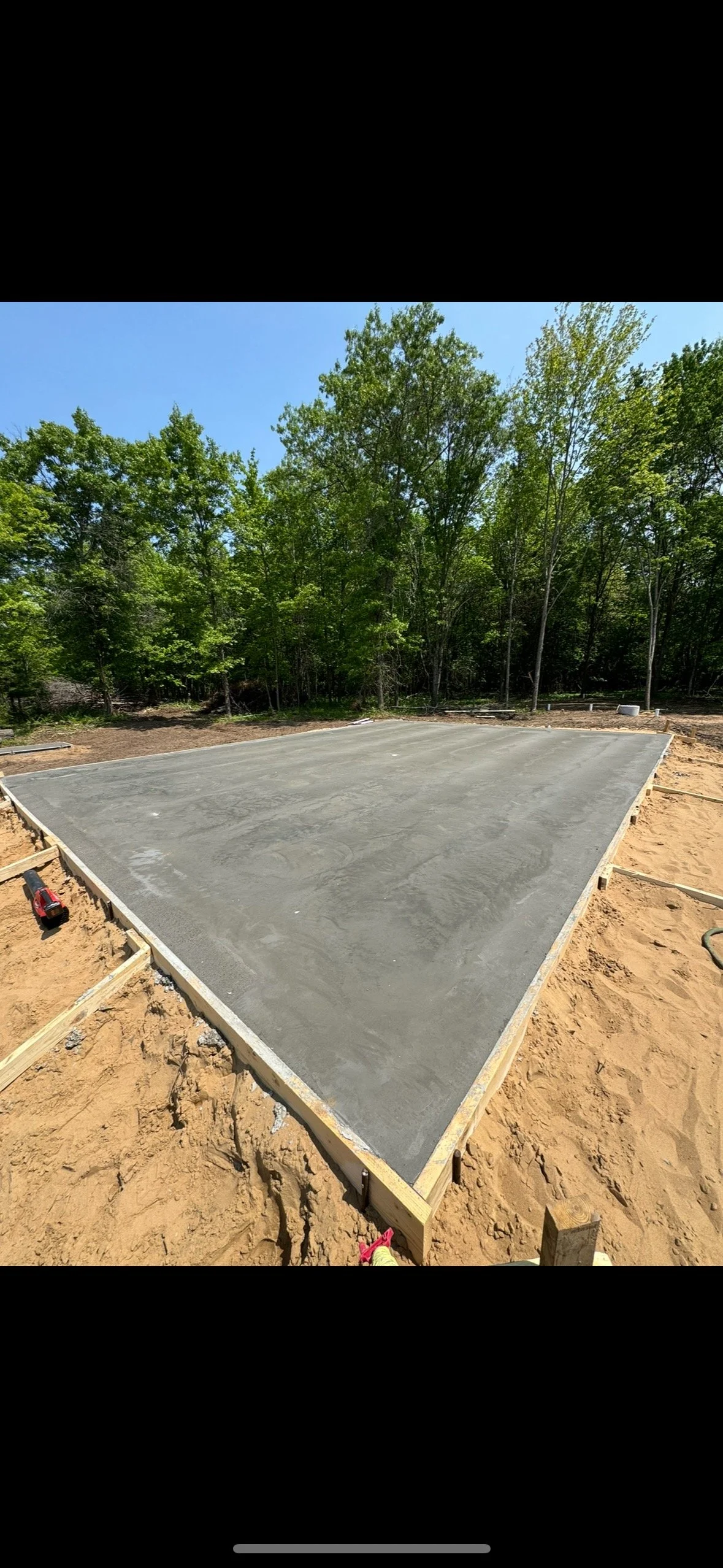 Freshly poured concrete slab on a construction site with wooden framing, surrounded by dirt and trees in the background under a clear blue sky.