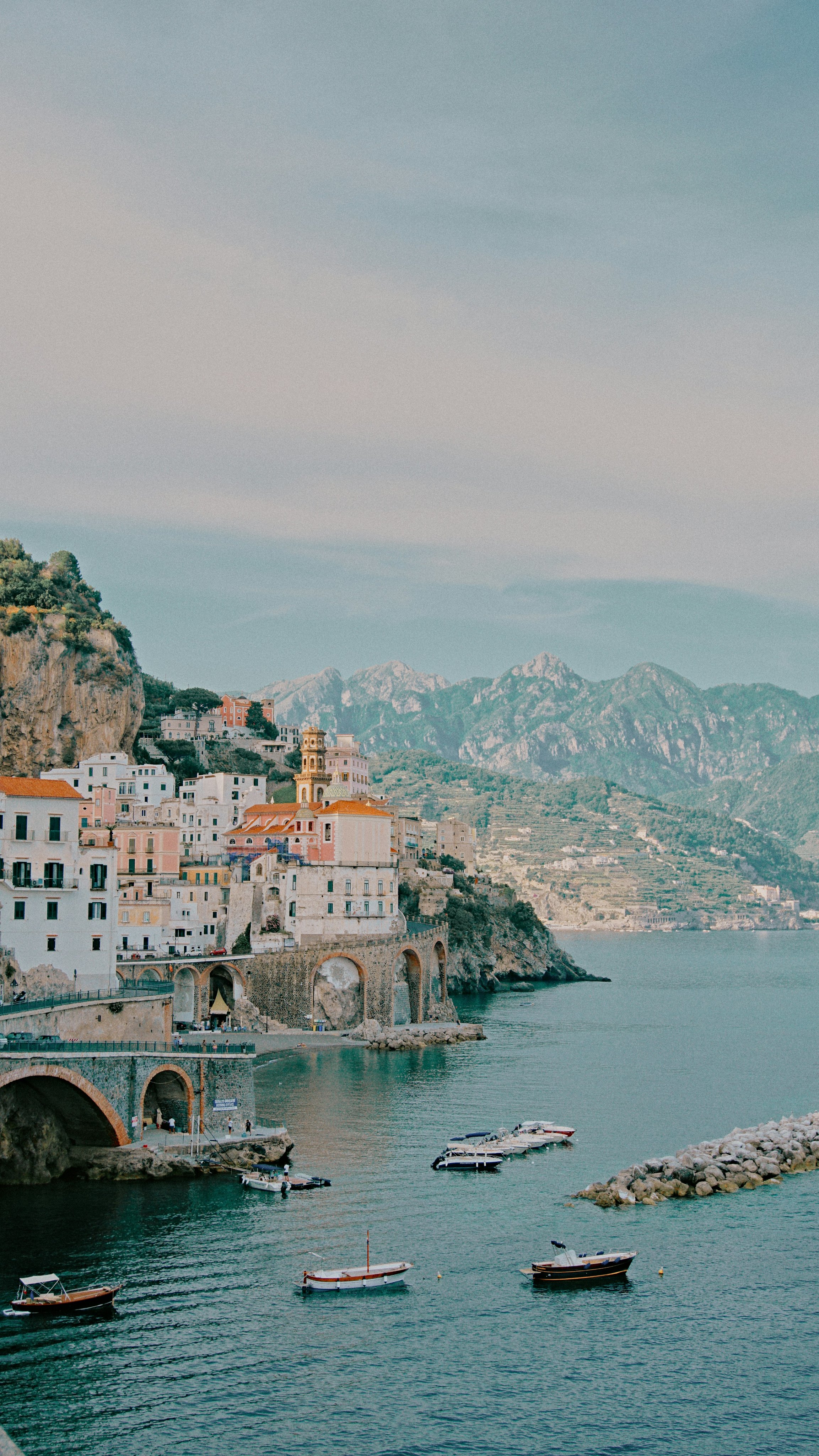 Coastal Italian village with colorful buildings on steep hillside overlooking the sea, boats docked in the water, mountains in the background.