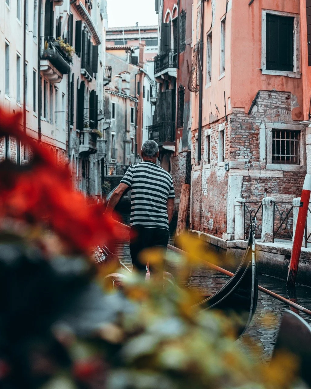 A man in a striped shirt riding a gondola through a narrow canal in Venice, Italy, with old buildings on either side.