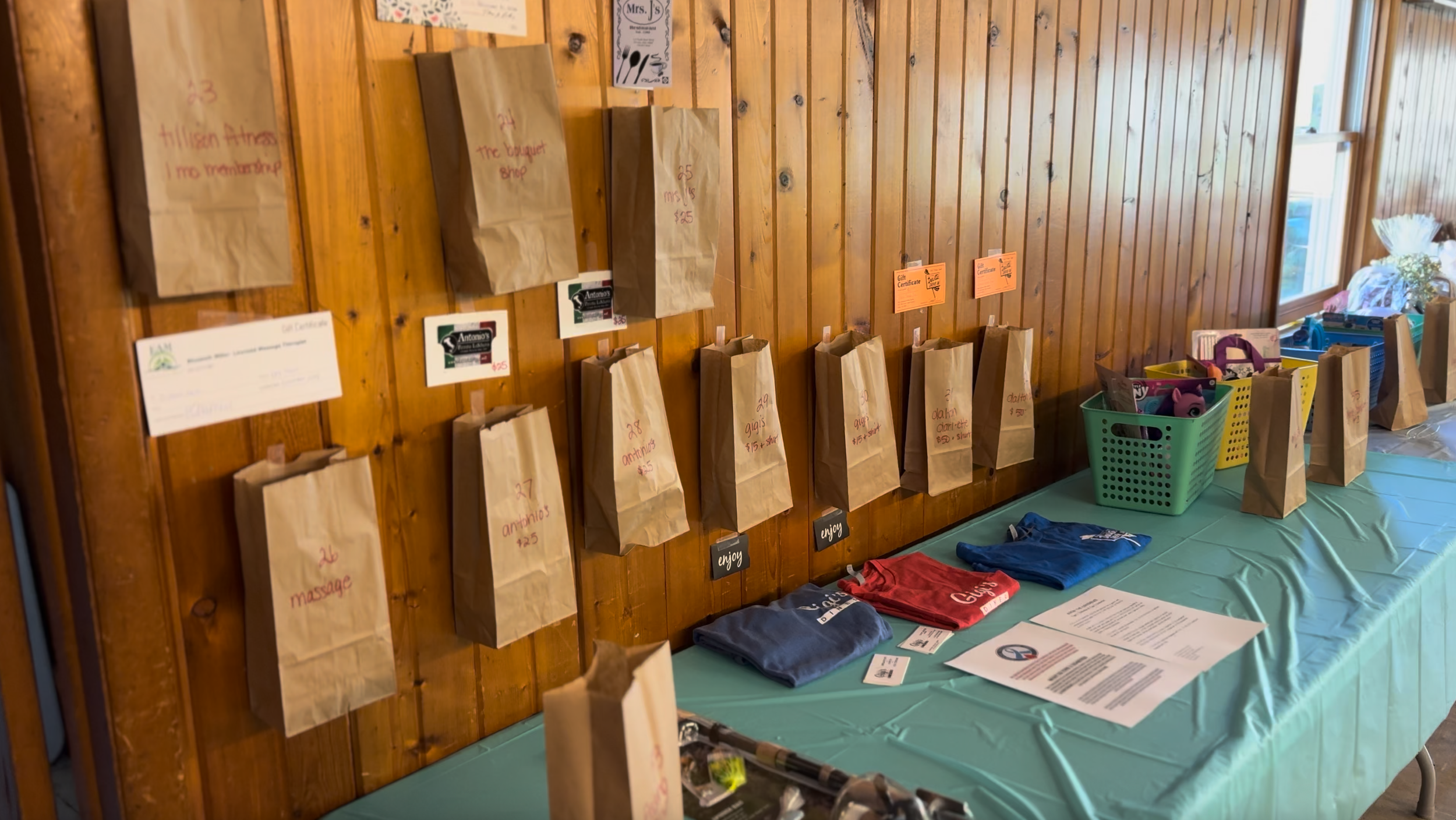 Fundraiser for type 1 diabetes support. A wooden wall with paper bags hanging, each labeled with different items and prices. A table covered with a green tablecloth holds folded T-shirts, baskets with more bags, and informational papers.