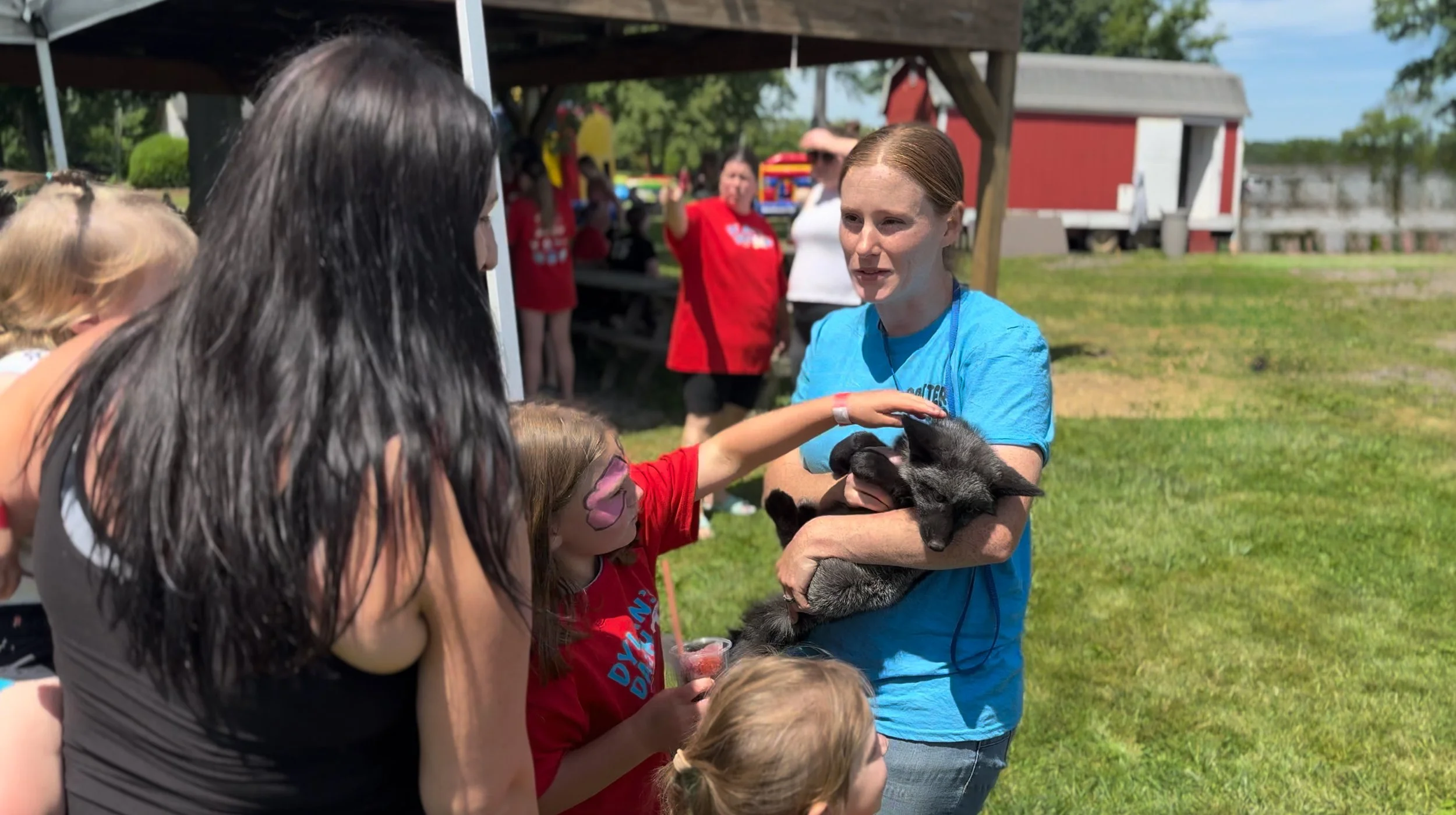 A woman holding a black baby fox in her arms as children and adults gather around outdoors on a sunny day, at a petting zoo for a fundraiser, with a red barn and green grass in the background.