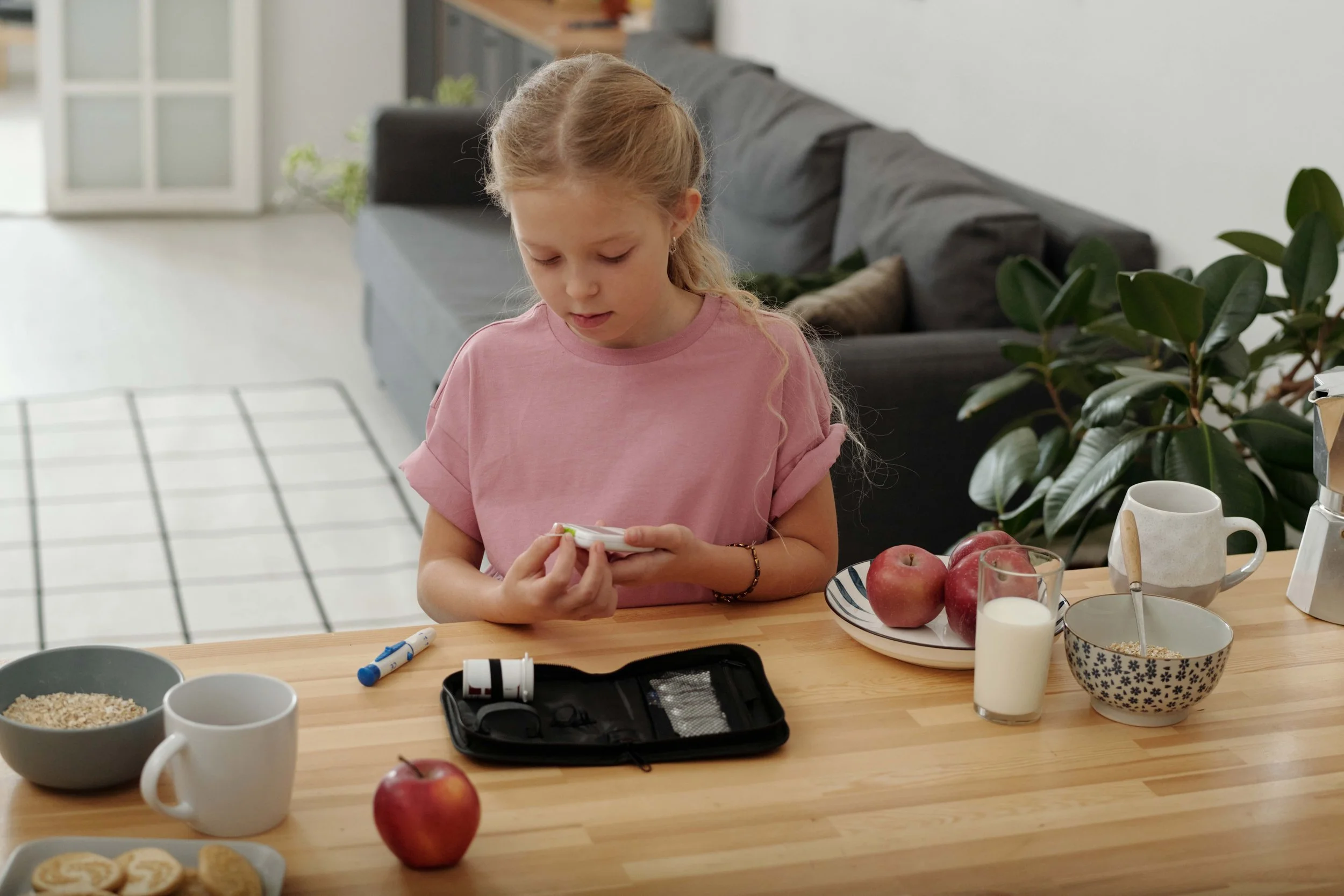 A young girl with long, curly blonde hair checking her blood sugar at a kitchen table.