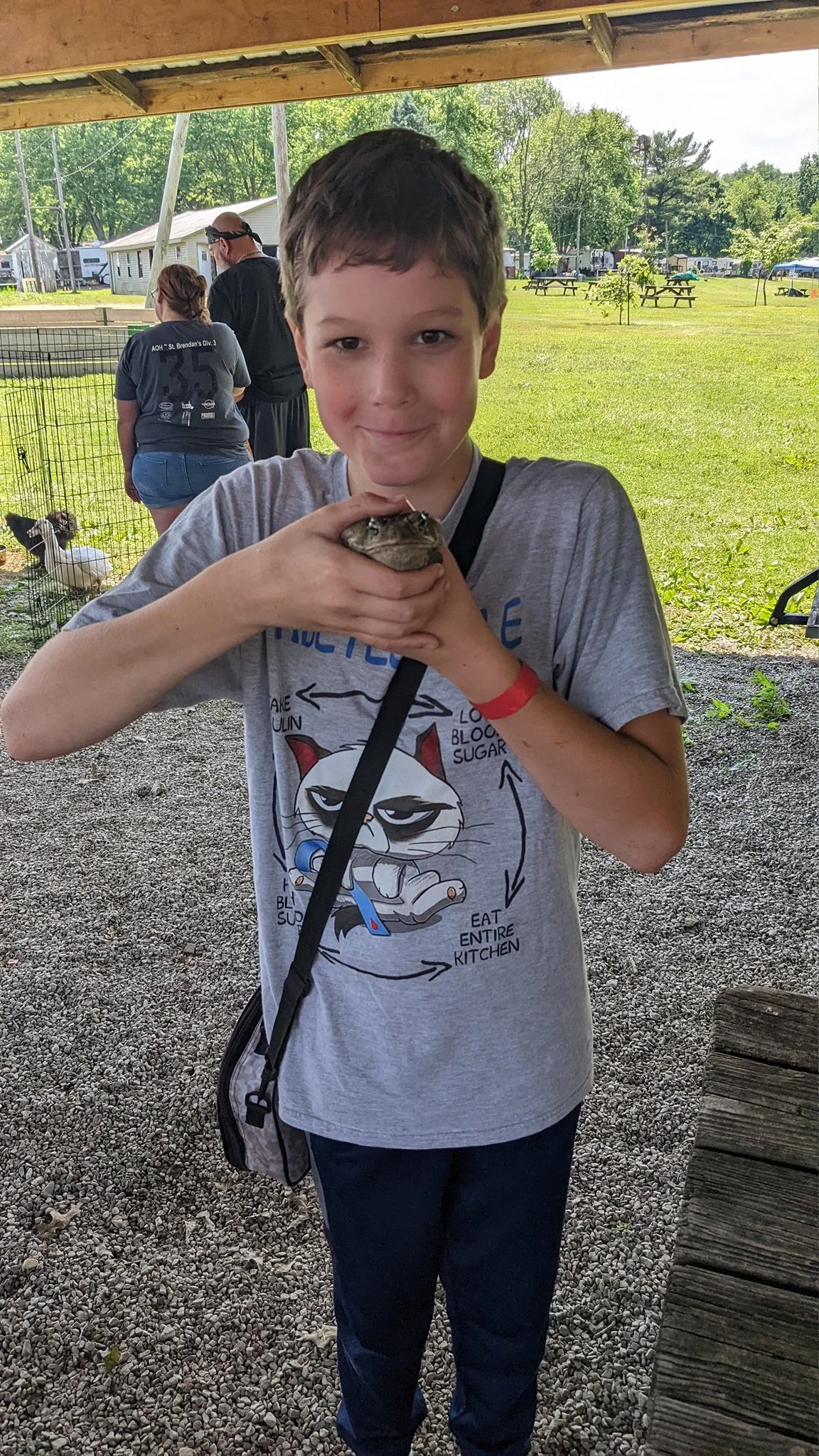 A young boy holding a large turtle at an outdoor event under a covered area with other people and animals in the background.