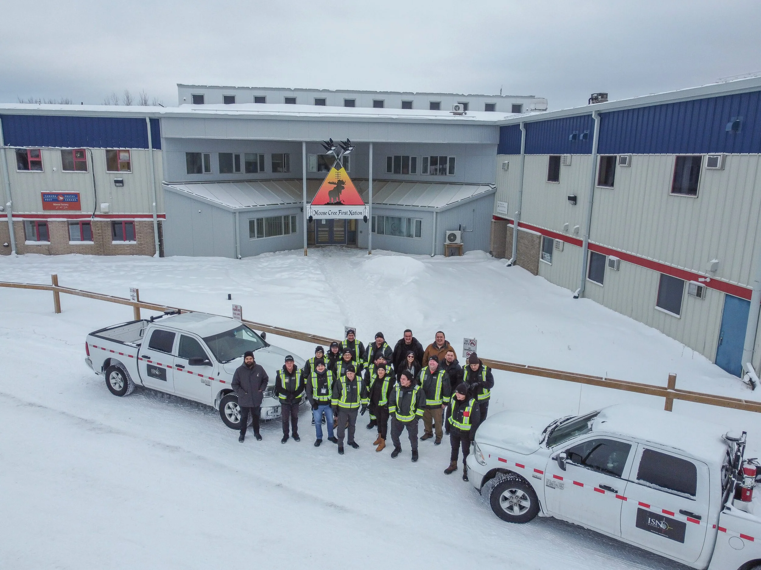 Group of people in winter clothing posing for a photo outdoors in snowy conditions, with two pickup trucks parked nearby, in front of a building with a Moose Cree First Nation sign.