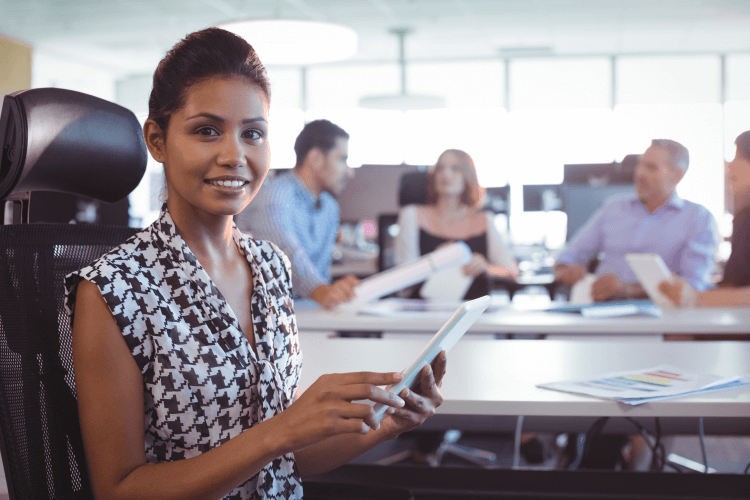 A woman sitting at a conference table in an office, holding a tablet, smiling at the camera with colleagues in the background.