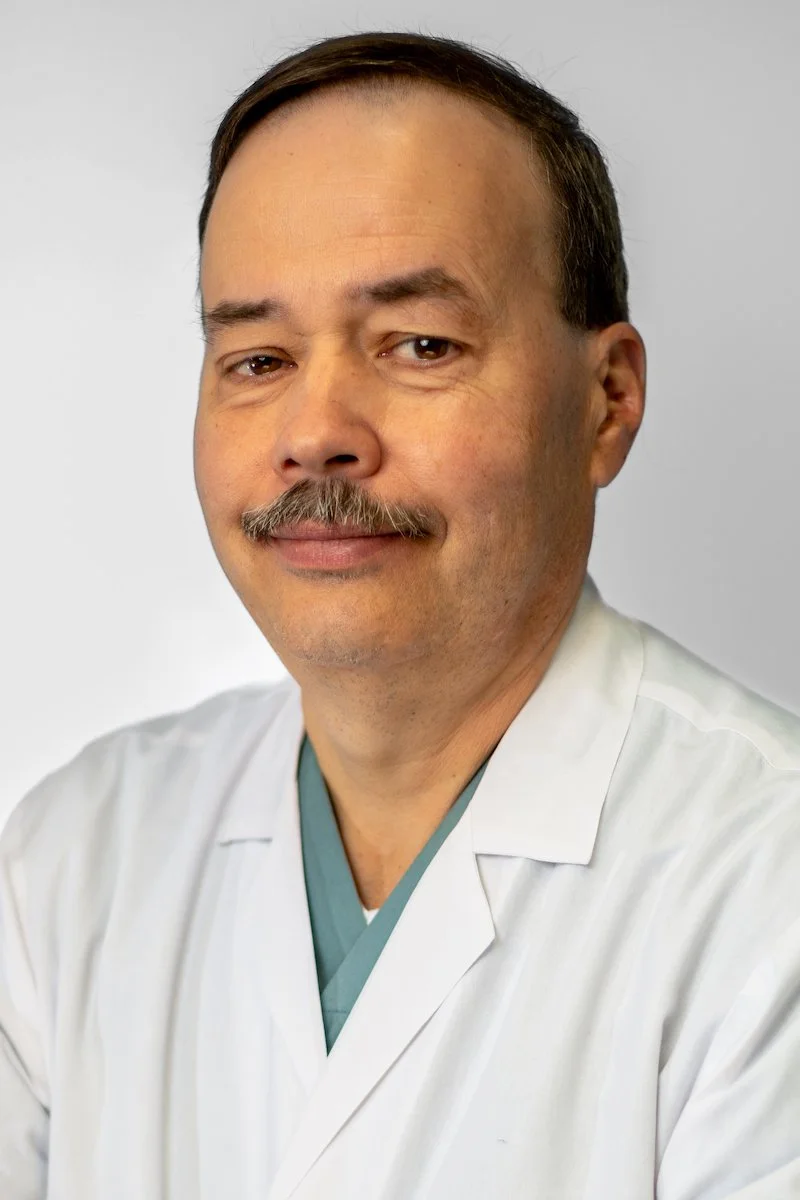 A man with a mustache wearing a white lab coat and medical scrubs, posing against a plain background.