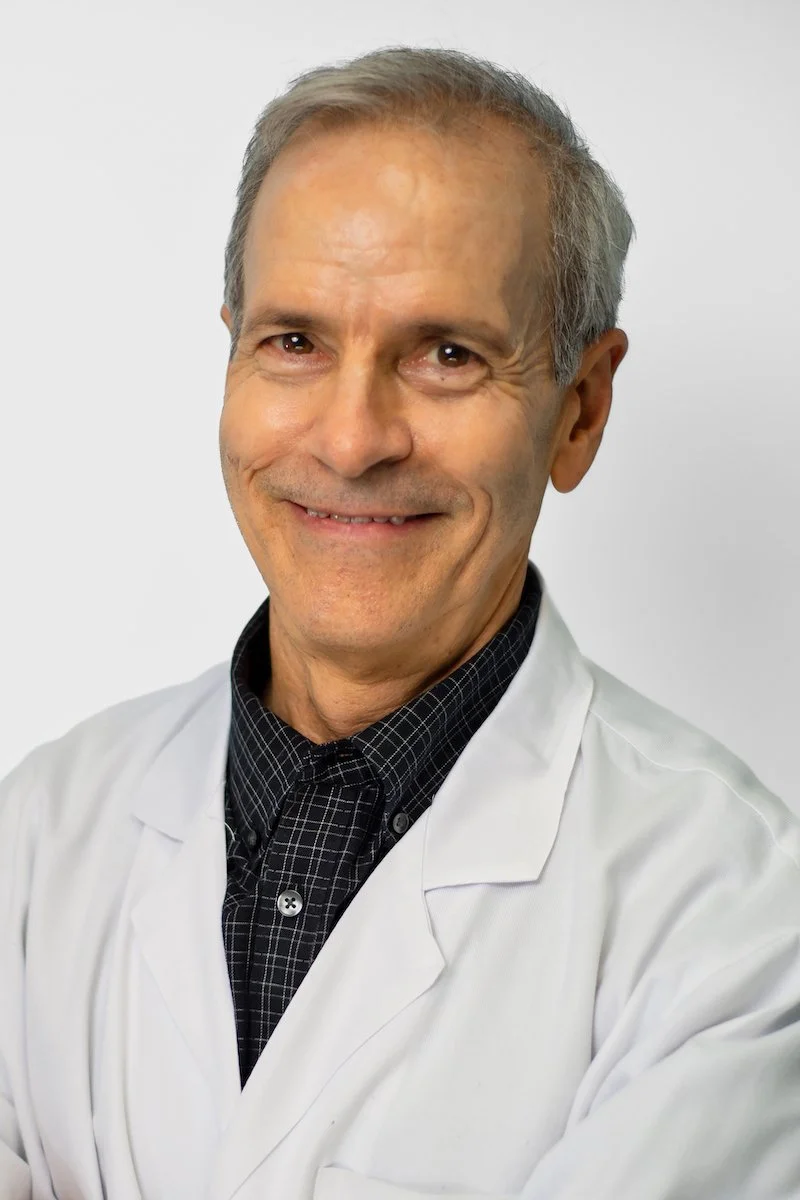 A middle-aged man with gray hair, wearing a white medical coat and a dark checkered shirt, smiling at the camera against a plain light background.
