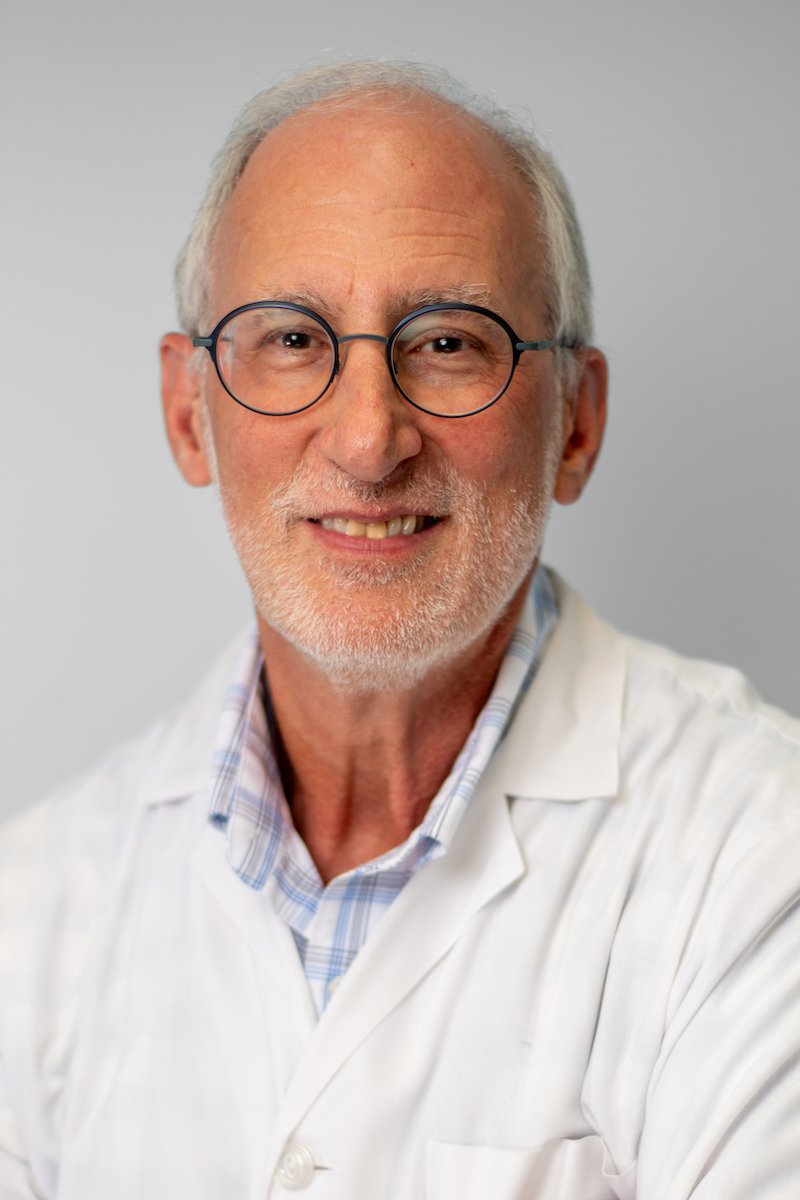 A smiling older man with glasses, white hair, and a beard, wearing a white lab coat over a plaid shirt, standing against a plain light gray background.