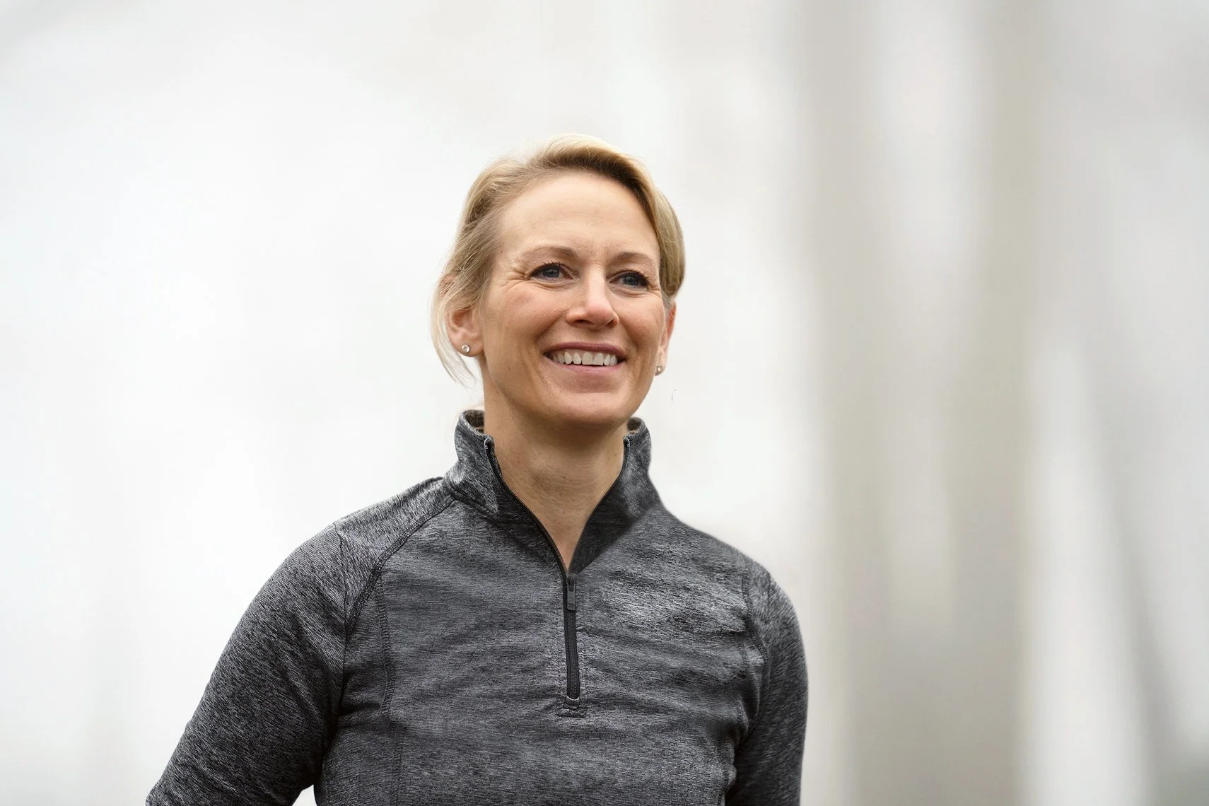 A smiling woman with short blonde hair wearing a dark gray athletic top with a quarter-zip, standing indoors against a plain, light-colored background.