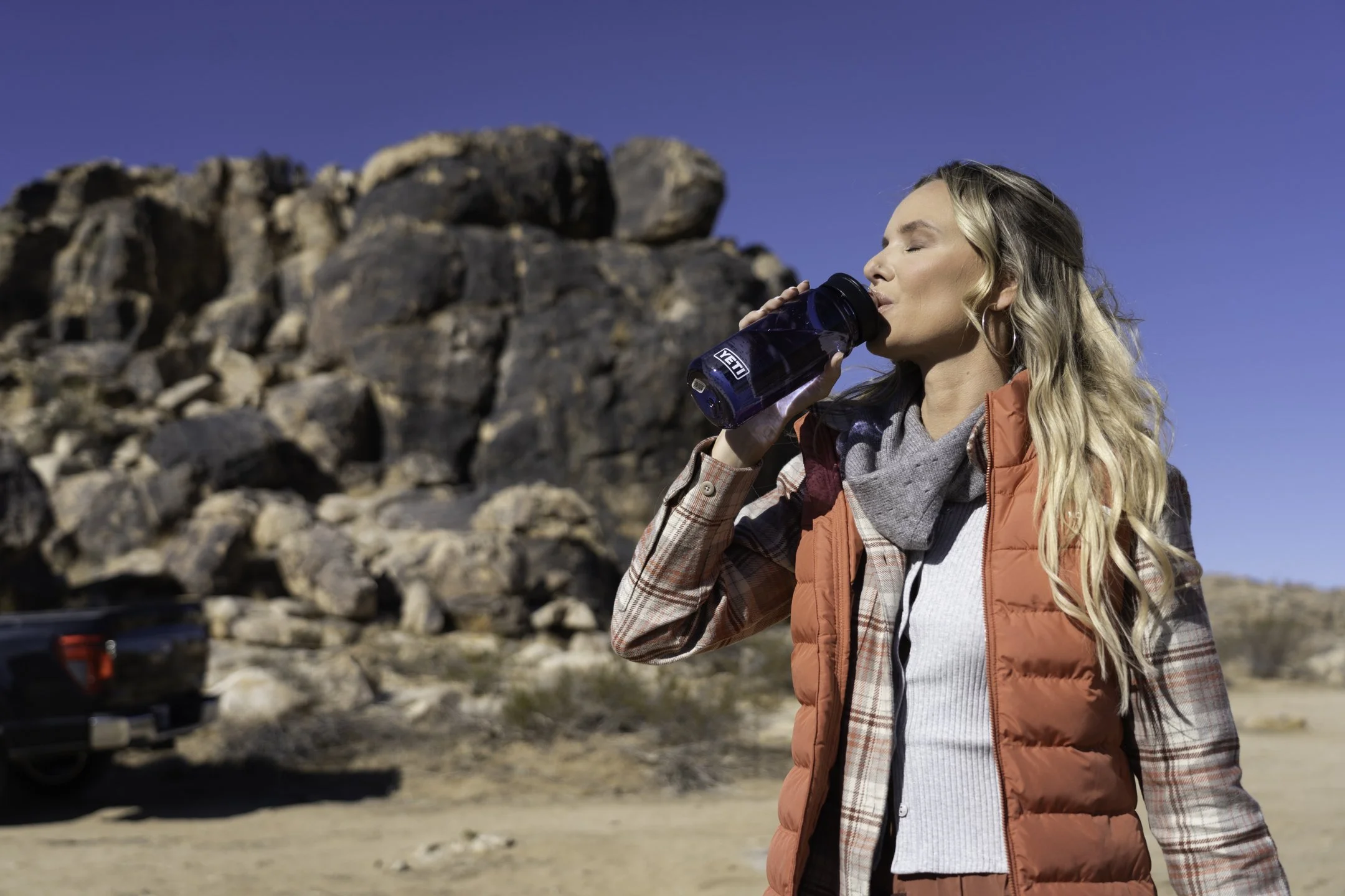 A woman with blonde hair wearing a red vest, plaid shirt, and gray scarf drinking from a blue YETI water bottle in a rocky desert landscape under a clear blue sky.