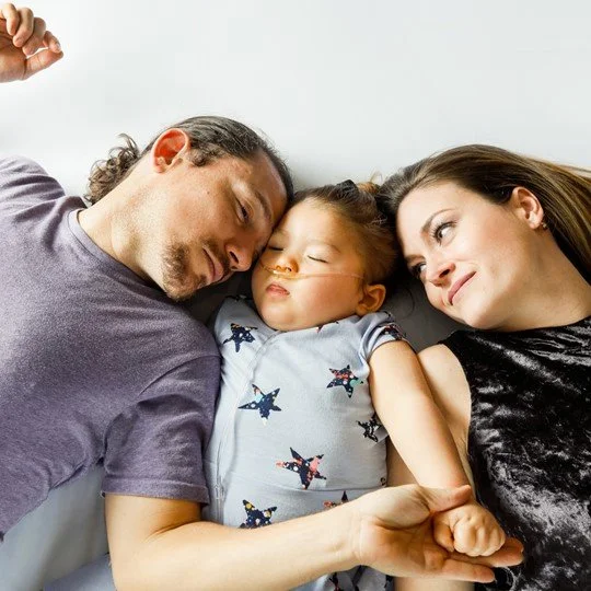 A family of three lying together on a bed, holding hands, with the father and mother smiling and the child sleeping peacefully.