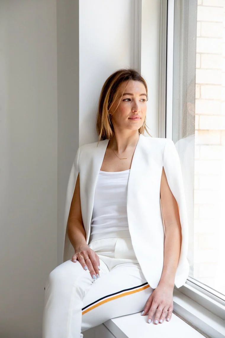 A woman sitting on a window sill, looking out the window with a contemplative expression. She is dressed in white and has light brown hair.